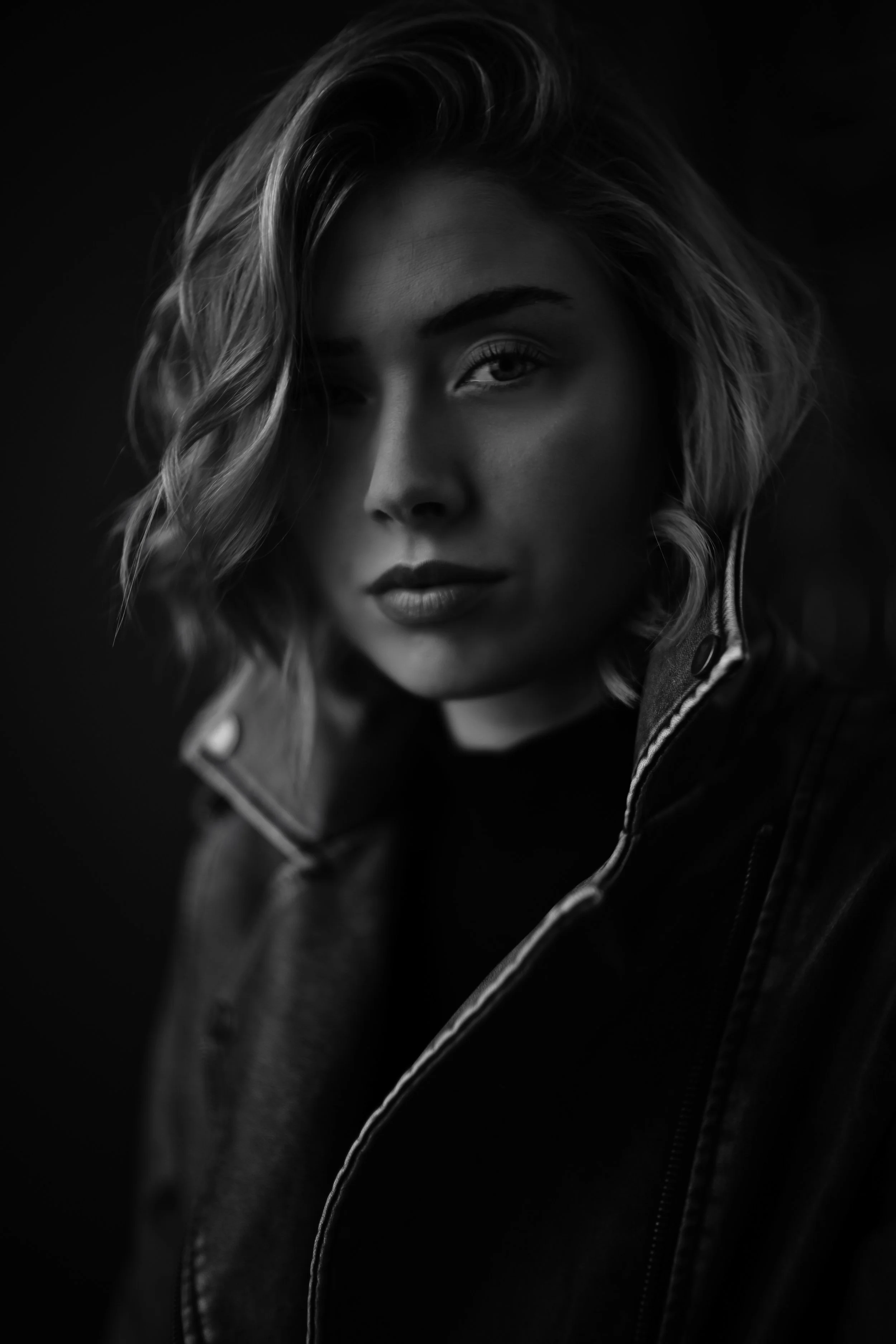 Schwarz-weiß-Fotografie eines Porträts einer jungen Frau mit welligem Haar, die eine Jacke trägt, vor dunklem Hintergrund.