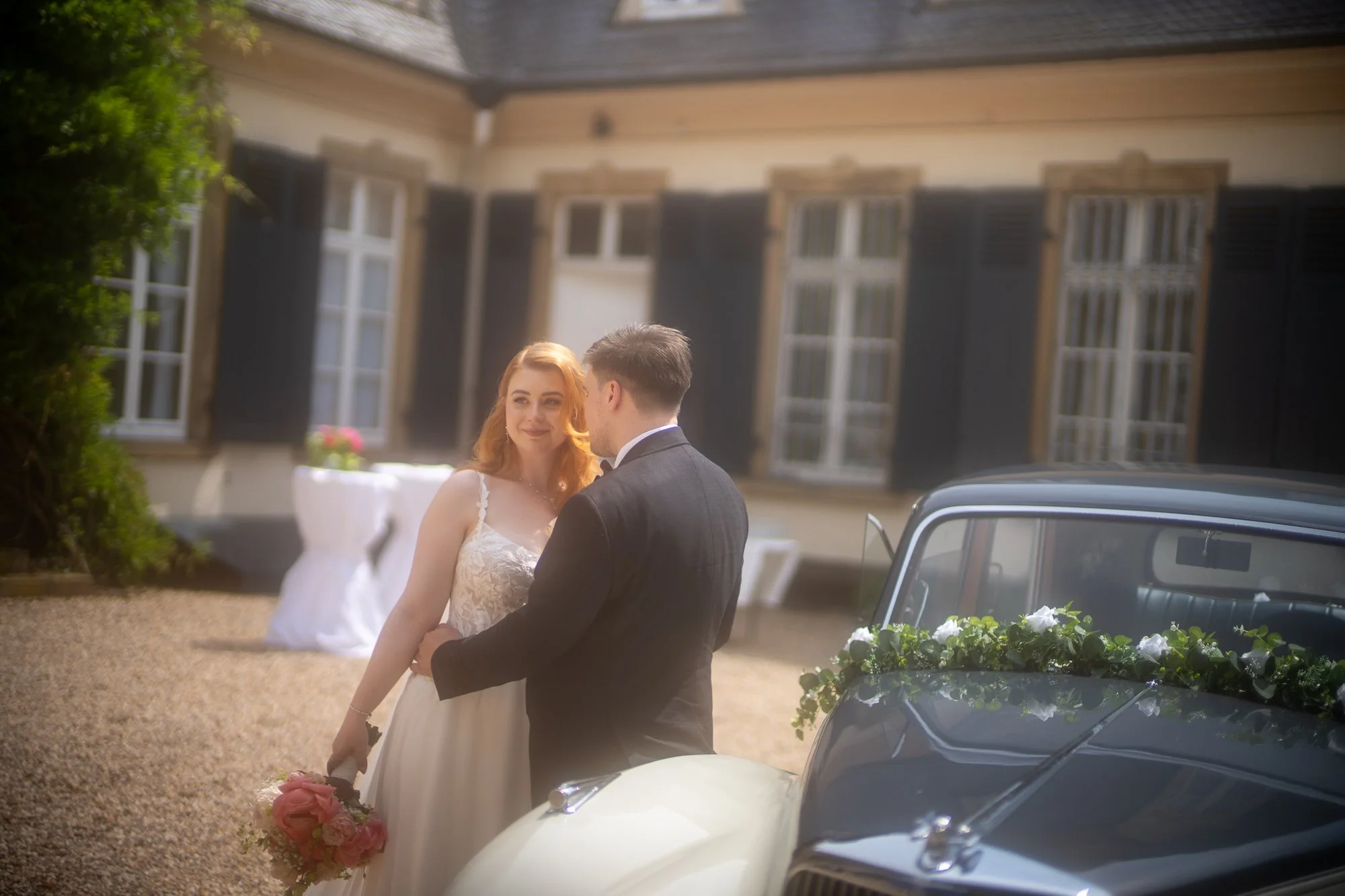 Verliebtes Brautpaar steht neben einem Oldtimer bei einer Hochzeit im Freien, vor einem Haus mit schwarzen Fensterläden.