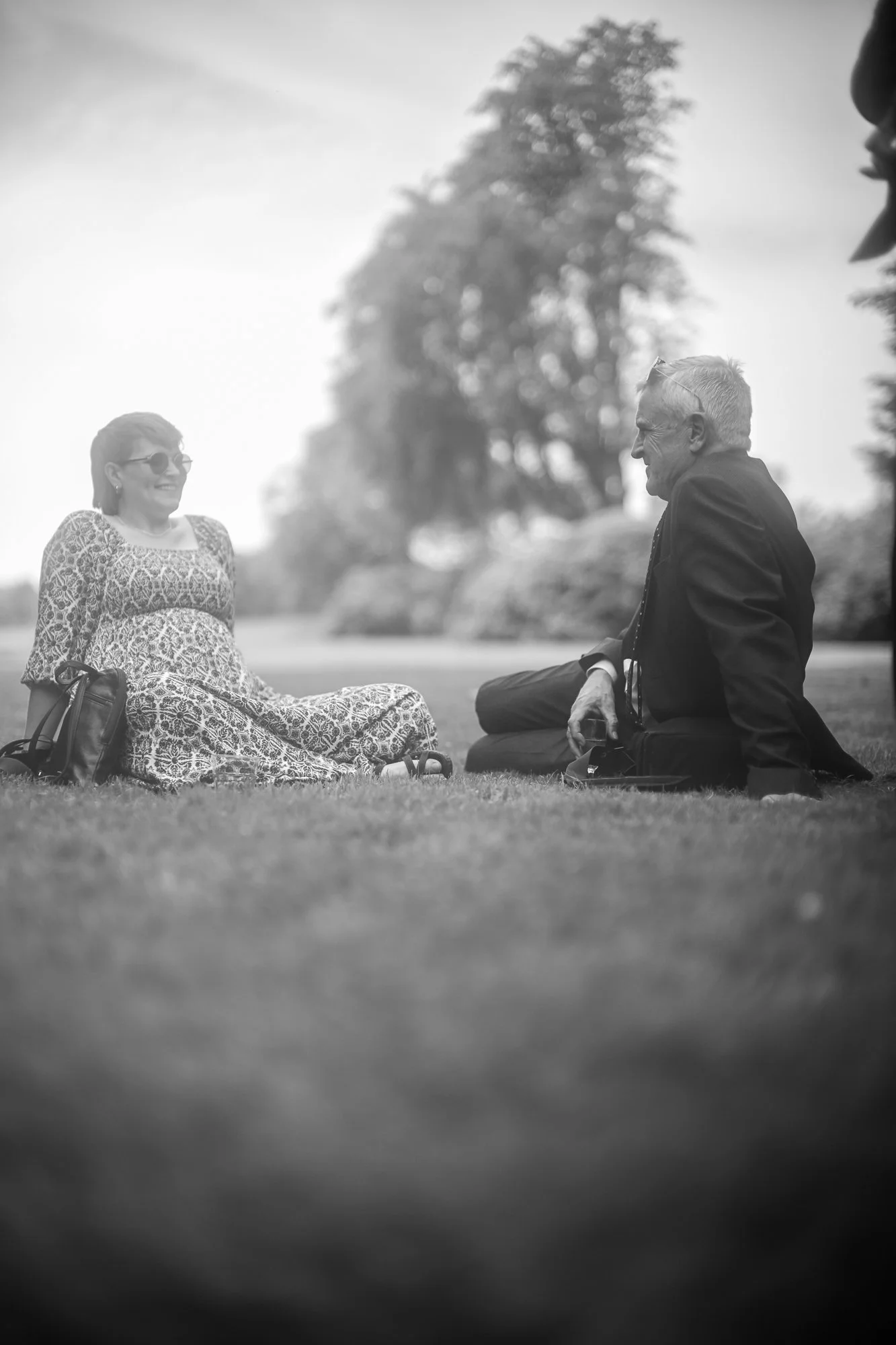 Ein älterer Mann und eine junge Frau sitzen auf dem Rasen im Park, lachen und führen ein Gespräch, die Frau trägt eine Sonnenbrille und ein langes Kleid, der Mann trägt einen Anzug, im Hintergrund sind Bäume und Himmel sichtbar.