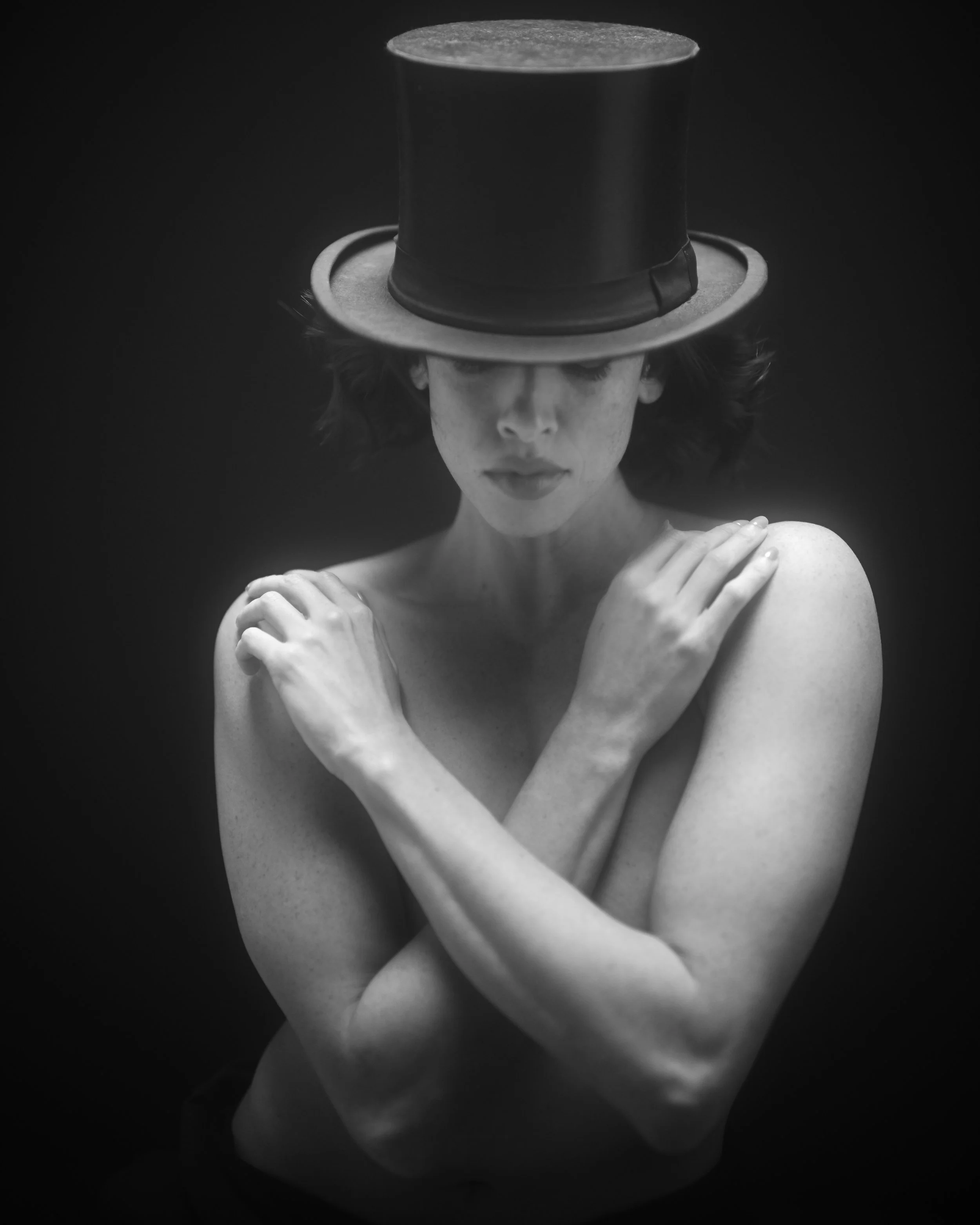 Schwarz-weiß-Foto einer Frau mit geschlossenen Augen und Gesichtsausdruck der Nachdenklichkeit, die einen schwarzen Zylinder-Hut trägt, der den oberen Teil ihres Kopfes bedeckt, und die Arme um ihre Schultern hält.