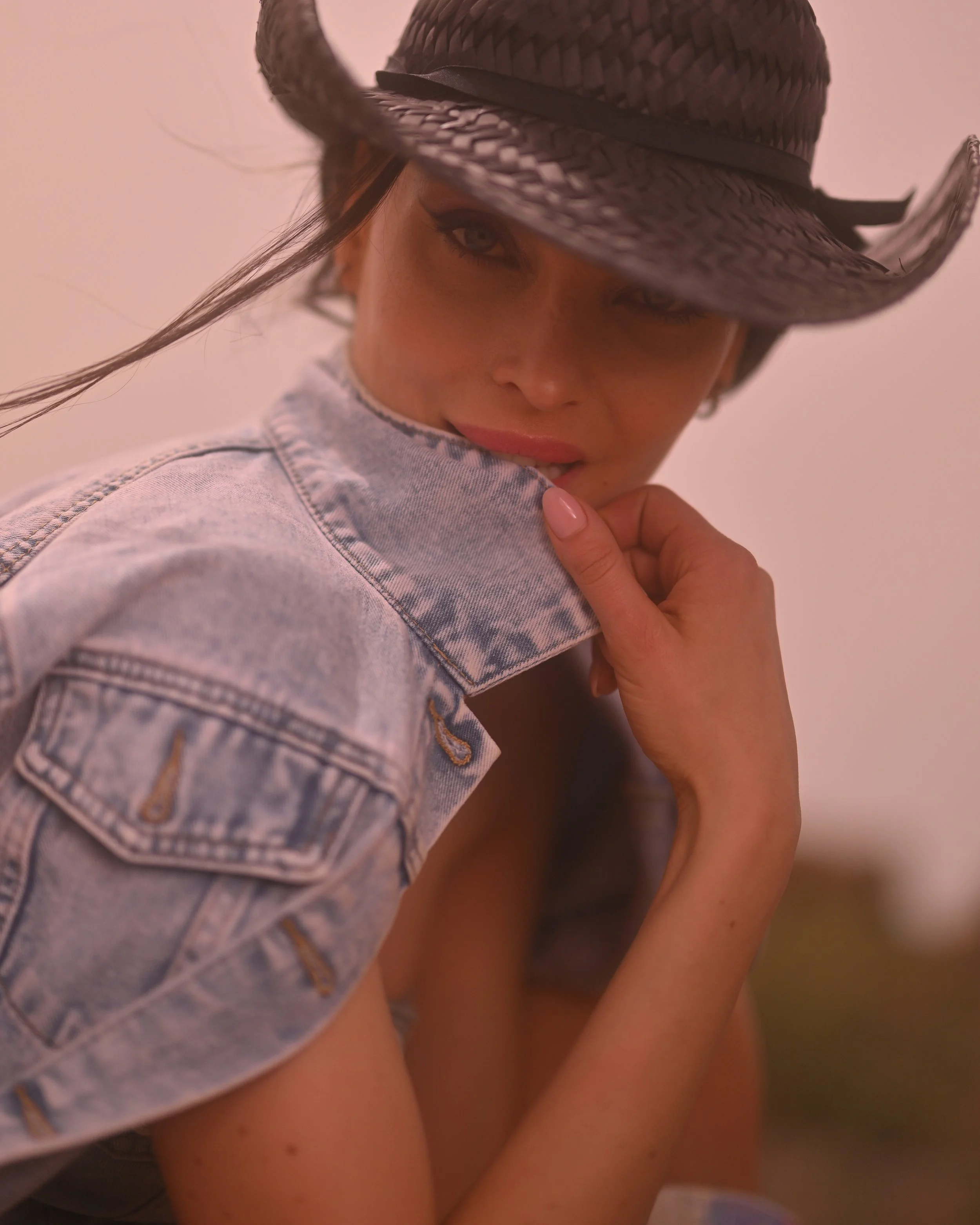 Nahaufnahme einer jungen Frau mit Hut, die sie sich leicht anlächelnd an den Hals fasst und direkt in die Kamera schaut.