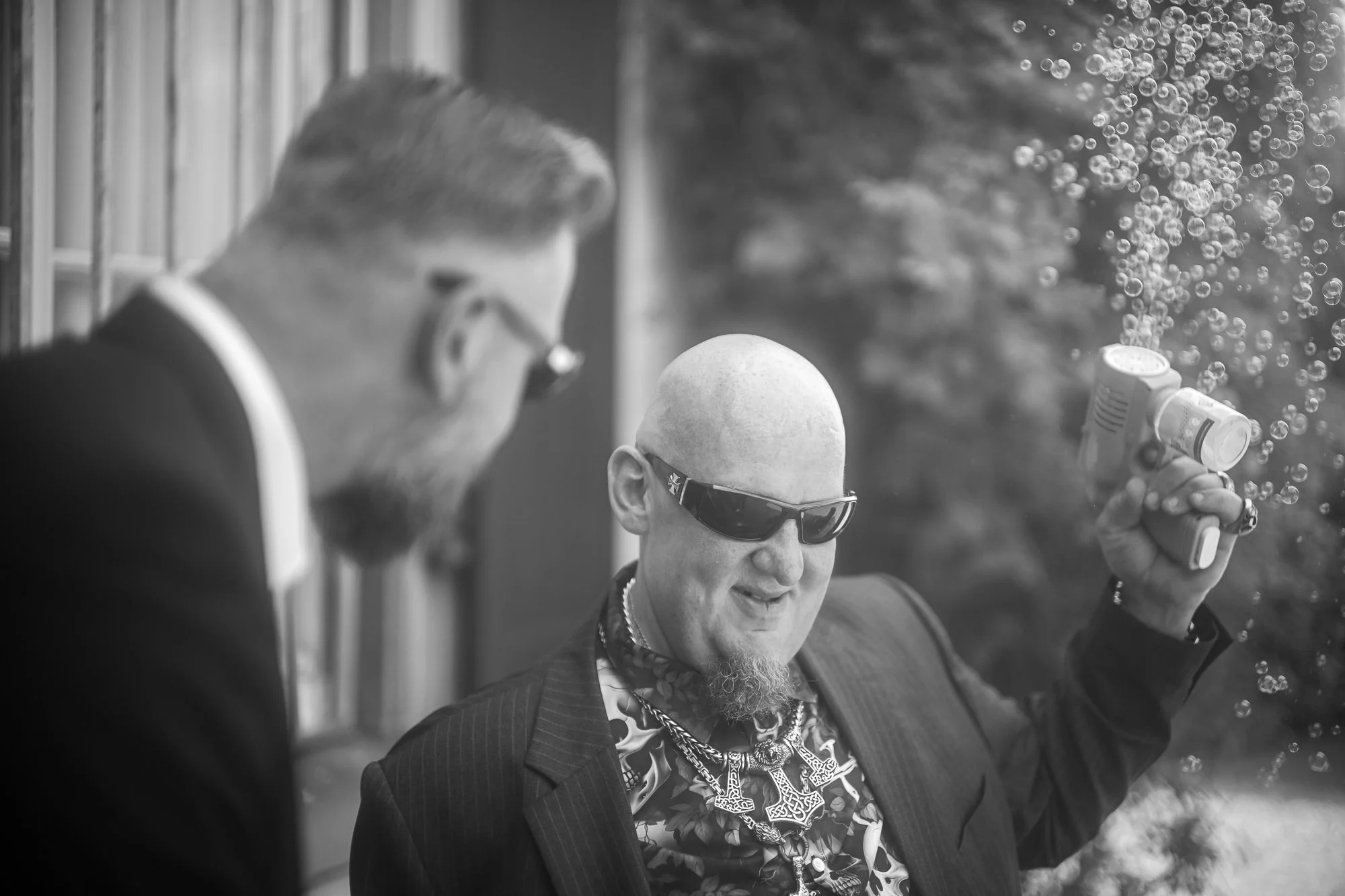 Zwei Männer, einer mit Brille, der andere mit Sonnenbrille, auf einem Balkon oder einer Terrasse, während einer mit einem Bubble Gun sprüht.