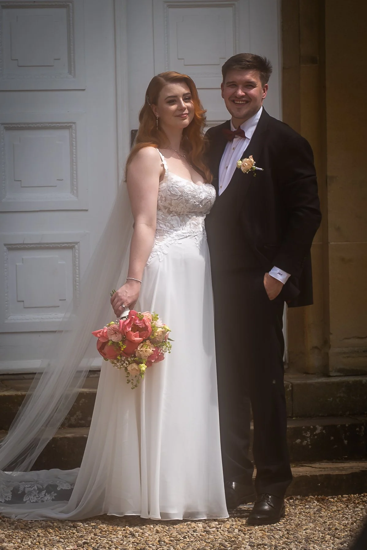 Ein Brautpaar bei einer Hochzeit, das Mädchen trägt ein weißes Brautkleid und hält einen Blumenstrauß, der Junge trägt einen schwarzen Anzug mit Fliege. Sie stehen vor einer weißen Tür und einem Steingarten.