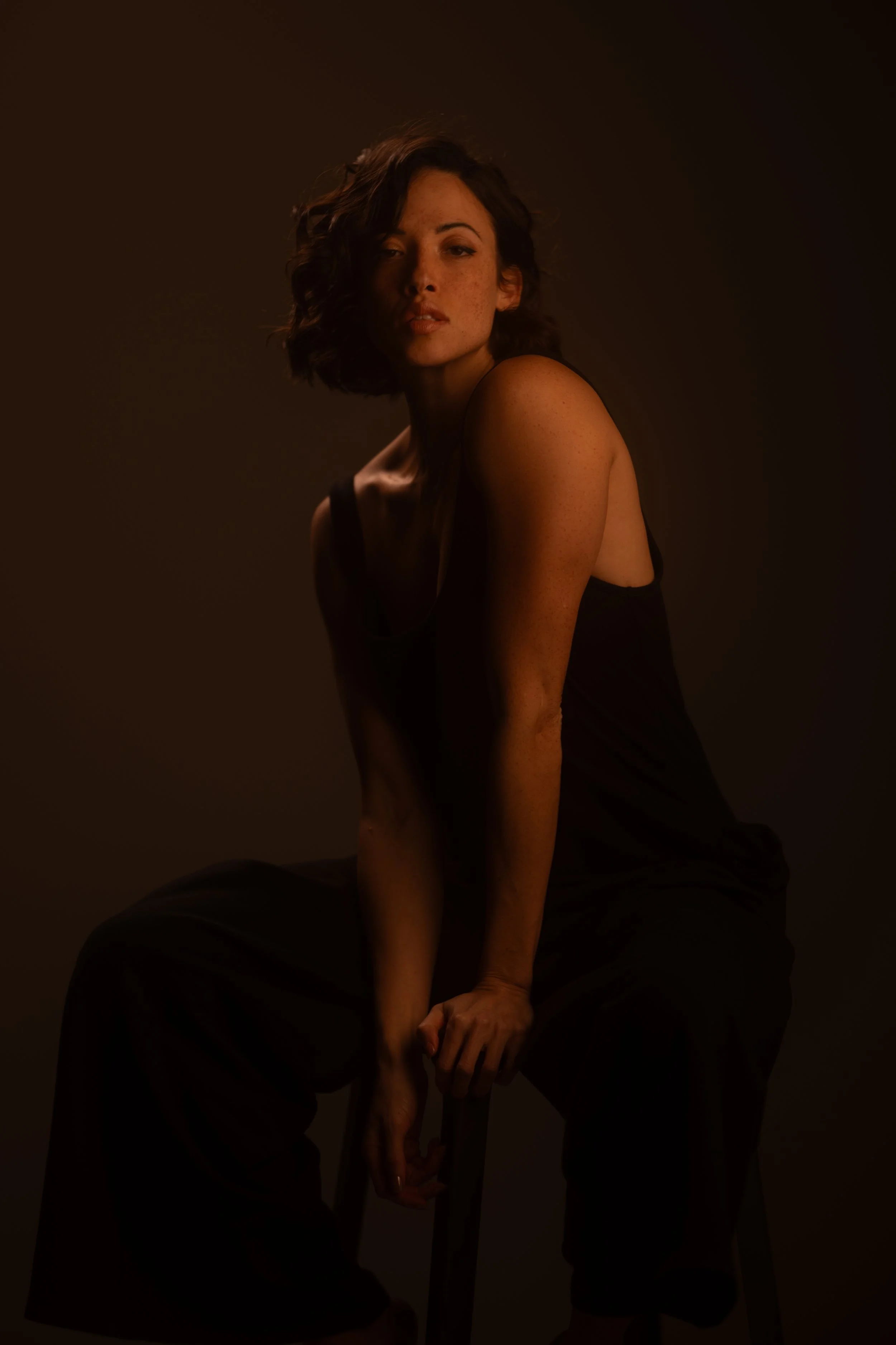 Ein junges Mädchen mit lockigen, dunklen Haaren sitzt auf einem Stuhl vor einem dunklen Hintergrund. Sie trägt ein schwarzes Kleid und schaut nachdenklich in die Kamera.