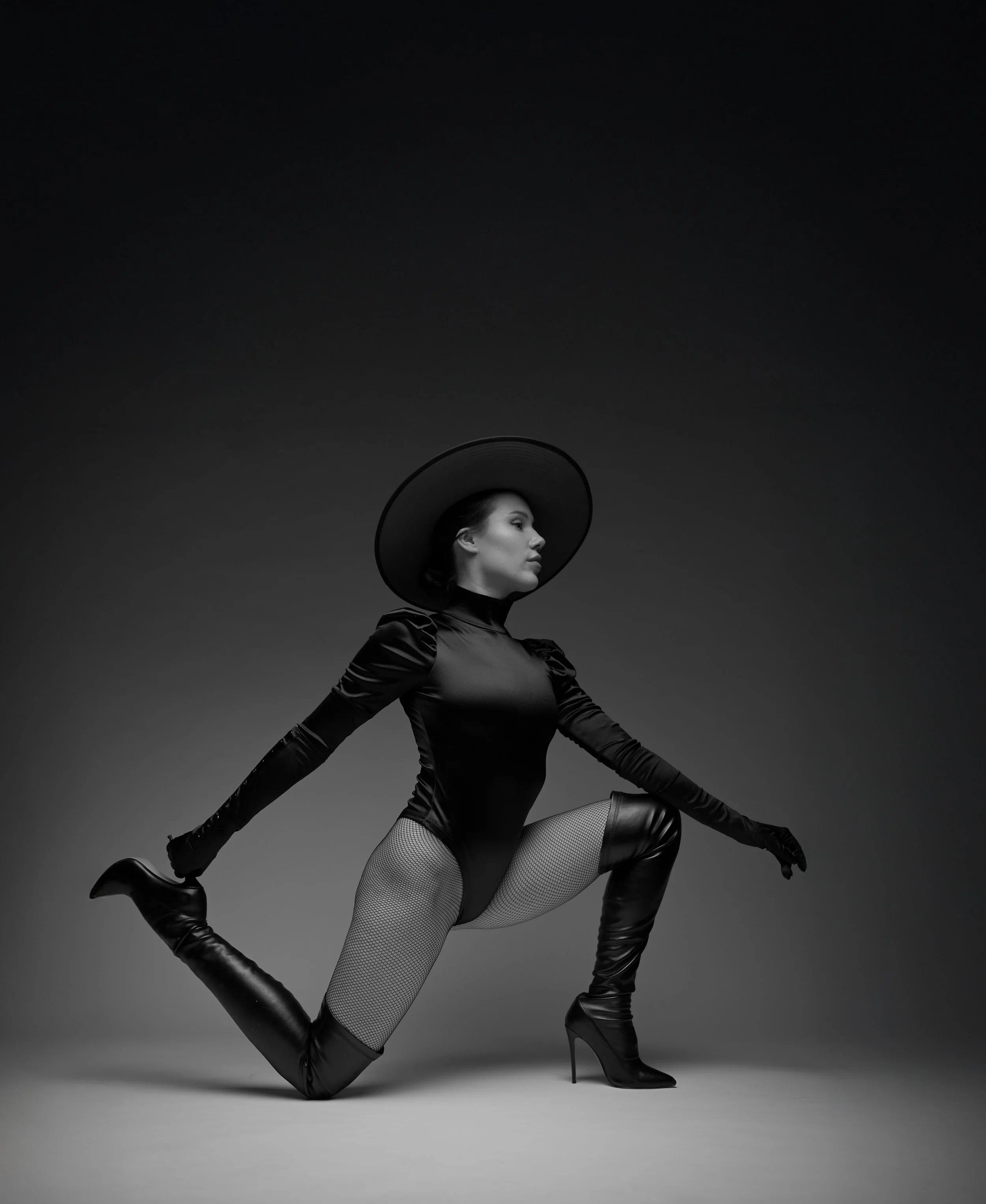Schwarze und weiße Mode- und Accessoire-Darstellung: Frau in einem schwarzen Kleid, hohen schwarzen Stiefeln, Netzstrümpfen, tragt einen großen schwarzen Hut, posiert in einer kreativen Haltung auf einer grauen Ebene, schwarz-weißer Hintergrund.