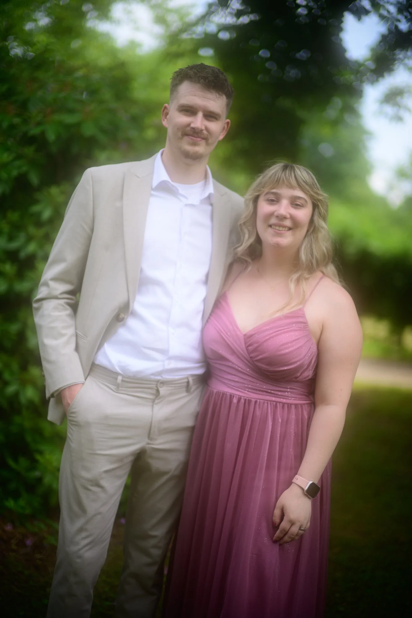 Ein Mann in einem beige Anzug und eine junge Frau in einem pinken Kleid stehen nebeneinander im Grünen, lächelnd in die Kamera.
