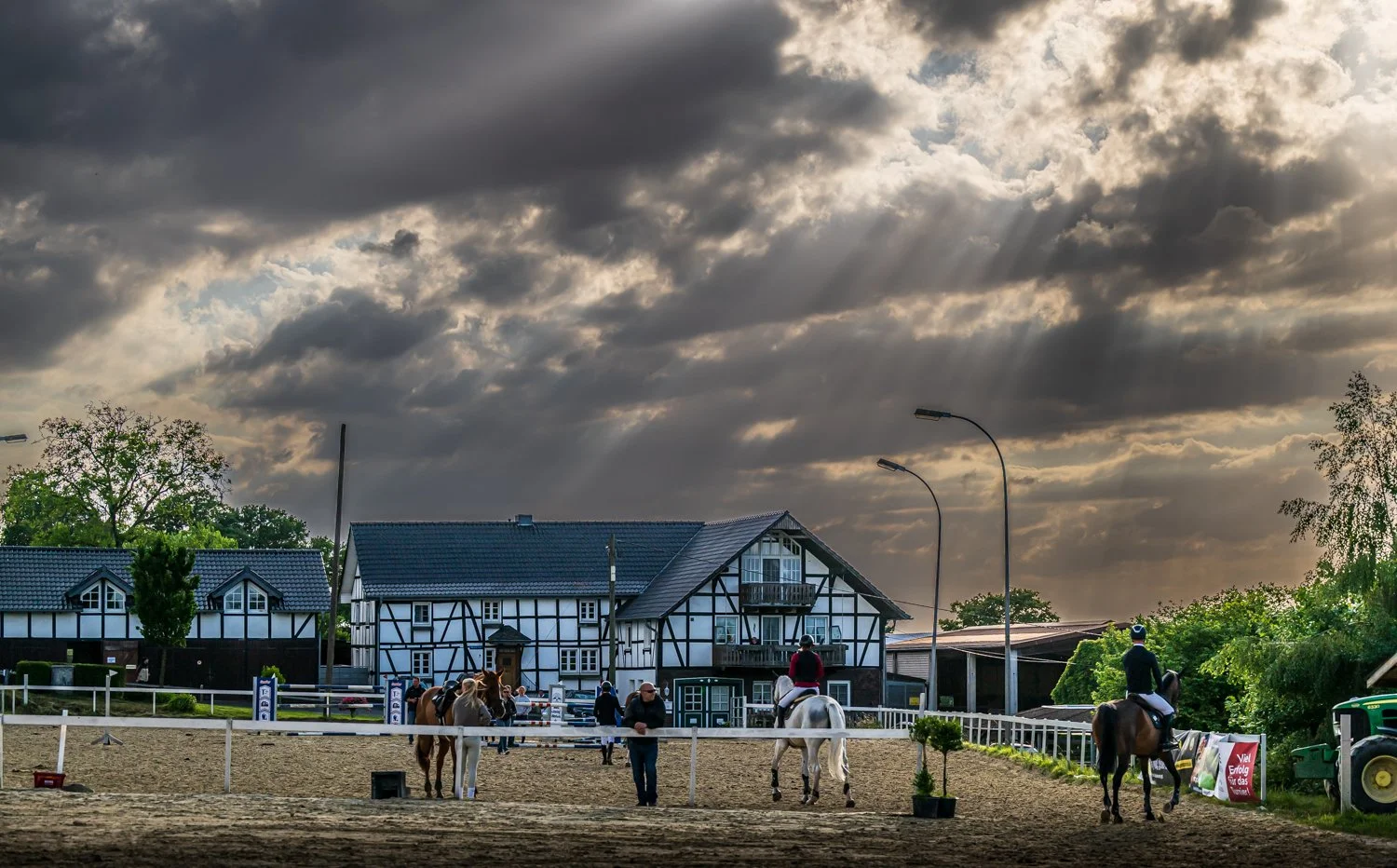 Reitturnier auf einem Reitplatz mit Pferden, Reitern, Betreuern und Zuschauern vor einem Fachwerkhaus bei bewölktem Himmel.