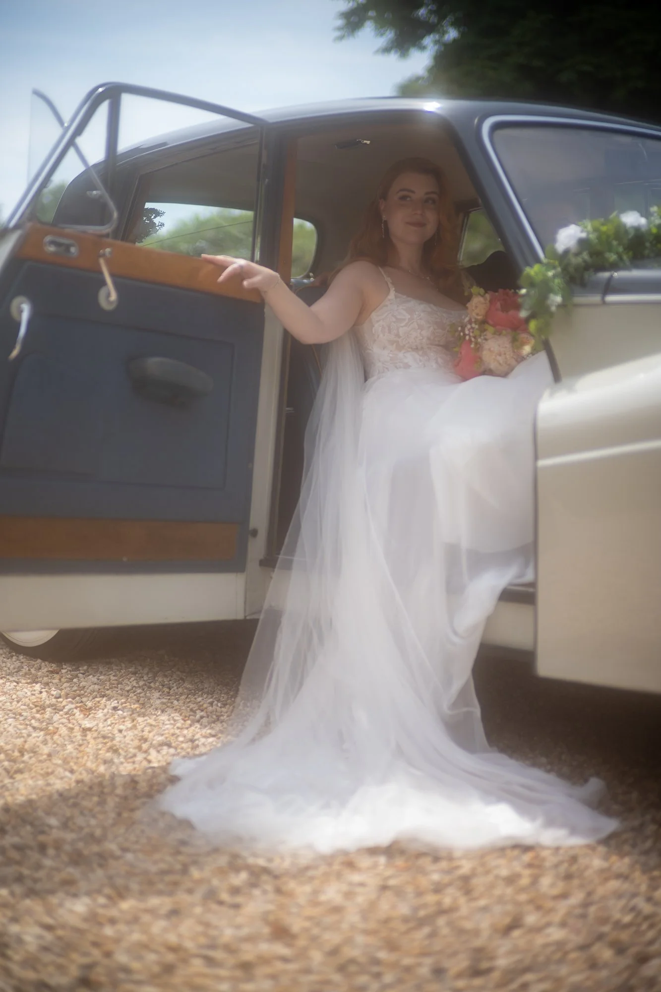 Eine Braut in einem weißen Hochzeitskleid sitzt in einem Oldtimer, hält einen Blumenstrauß und lächelt in die Kamera hinaus.