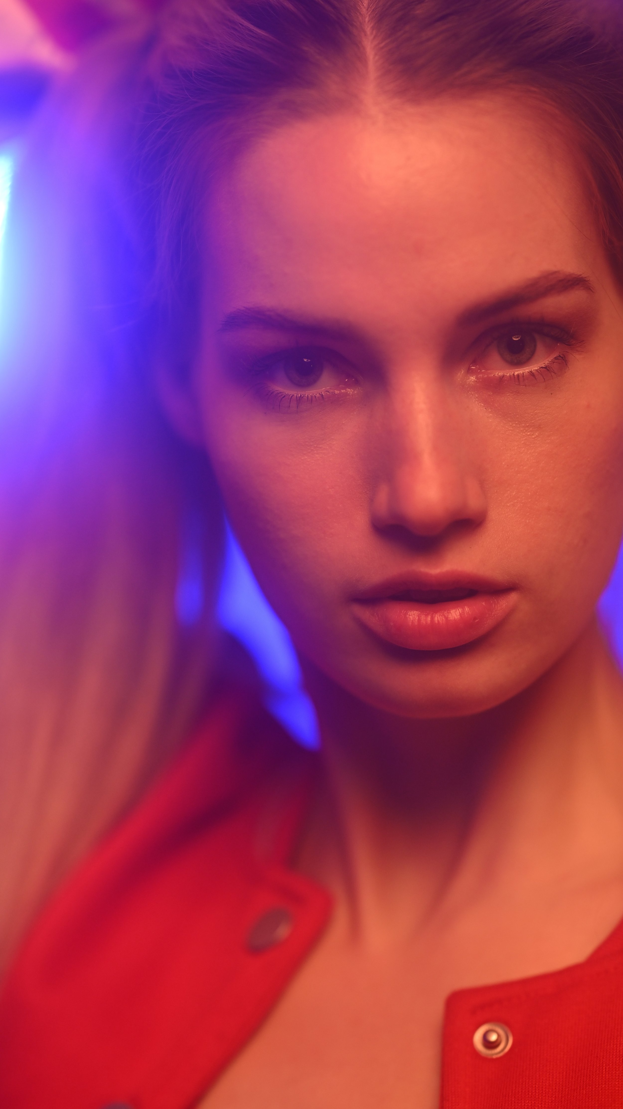 Nahaufnahme eines Menschen mit hellem Teint und braunen Augen, rote Haare, trägt eine rote Jacke, sichtbar im Vordergrund, im Hintergrund verschwommene bunte Lichter in Blau und Violett.