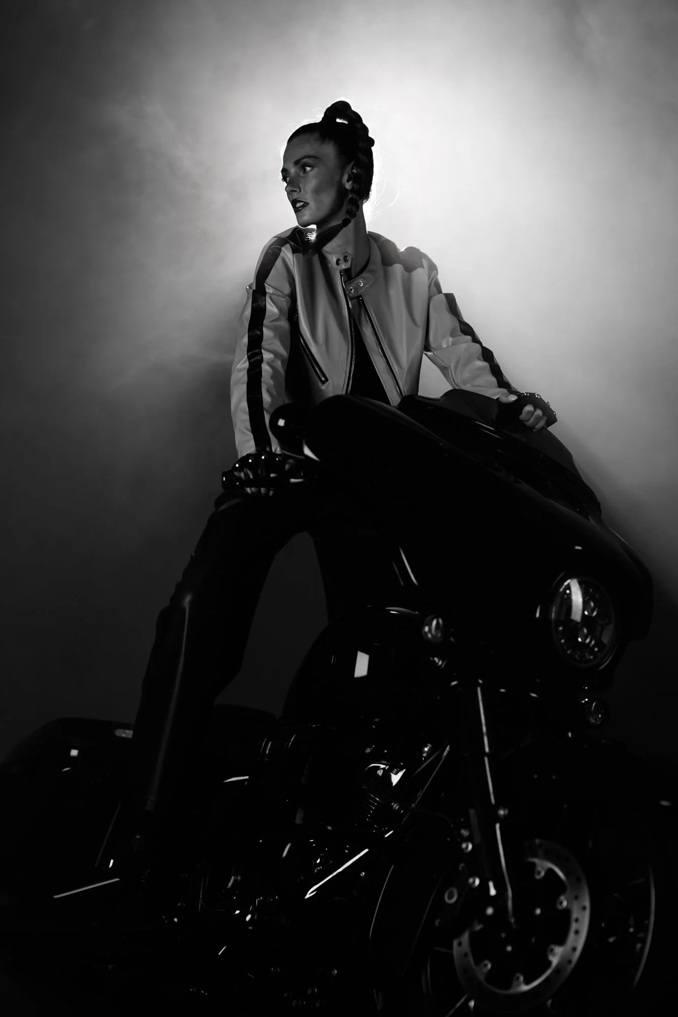 Schwarz-Weiß-Fotografie einer Frau mit einem Motorrad. Die Frau trägt eine Jacke und schaut zur Seite. Der Hintergrund ist hell, während das Motorrad dunkel ist.