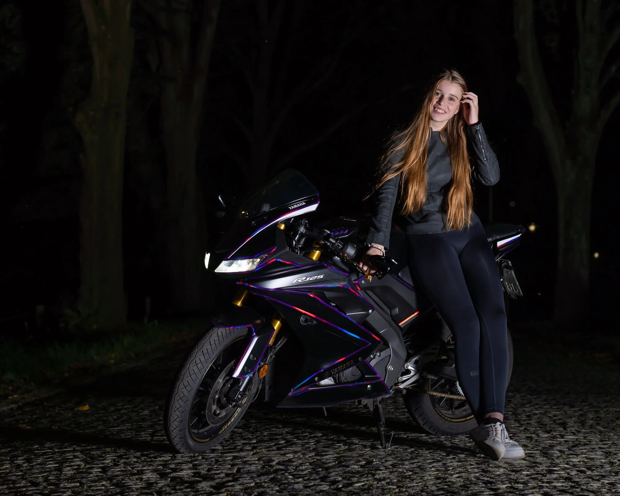 Junge Frau sitzt auf einer schwarzen Yamaha R125 Motorrad bei Nacht im Park, umgeben von Bäumen, lächelnd, trägt schwarze Jacke und Schuhe.