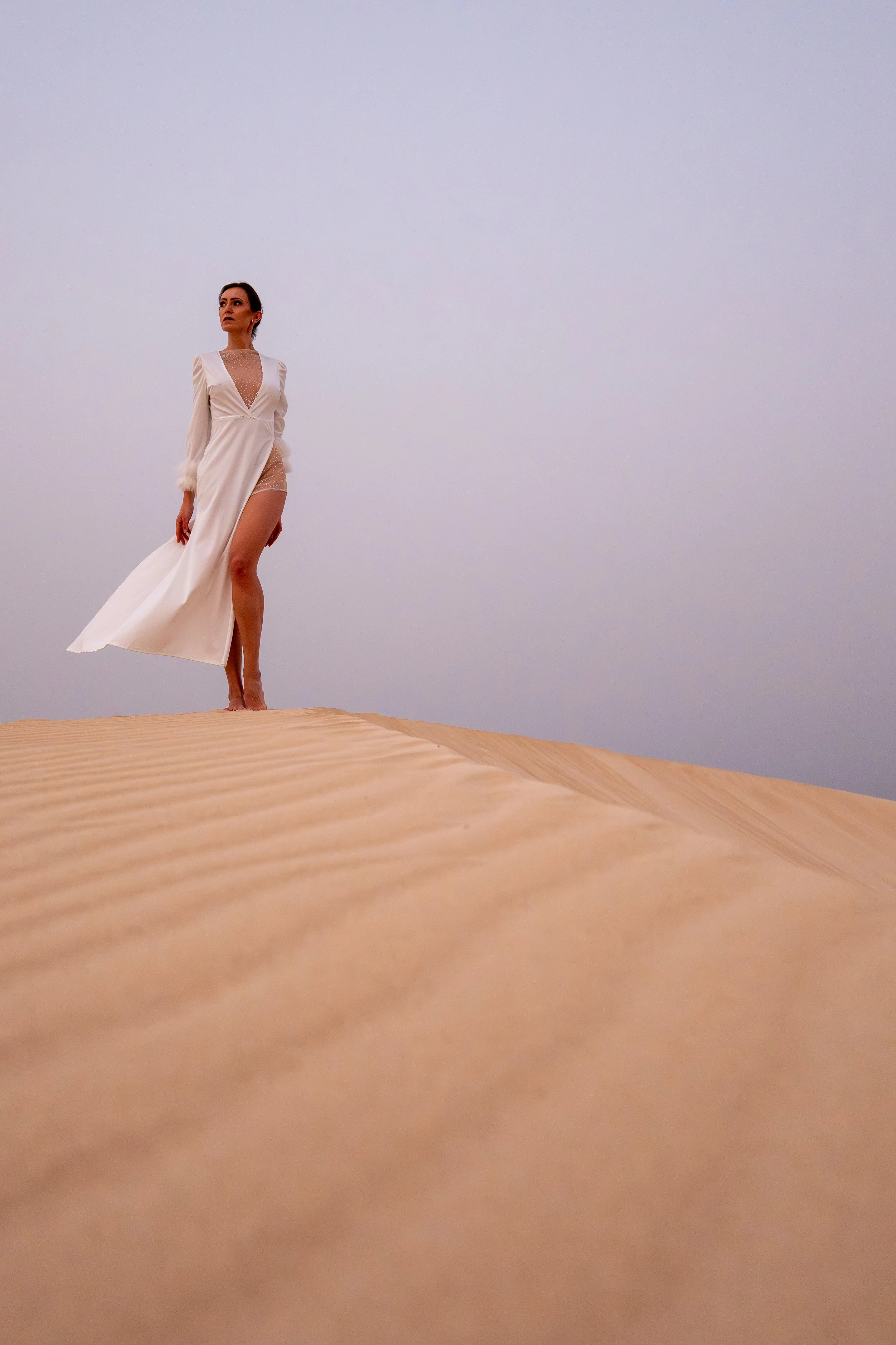 Frau in eleganter weißer Kleidung steht auf Sanddünen, blickt in die Ferne, overcast Himmel im Hintergrund.