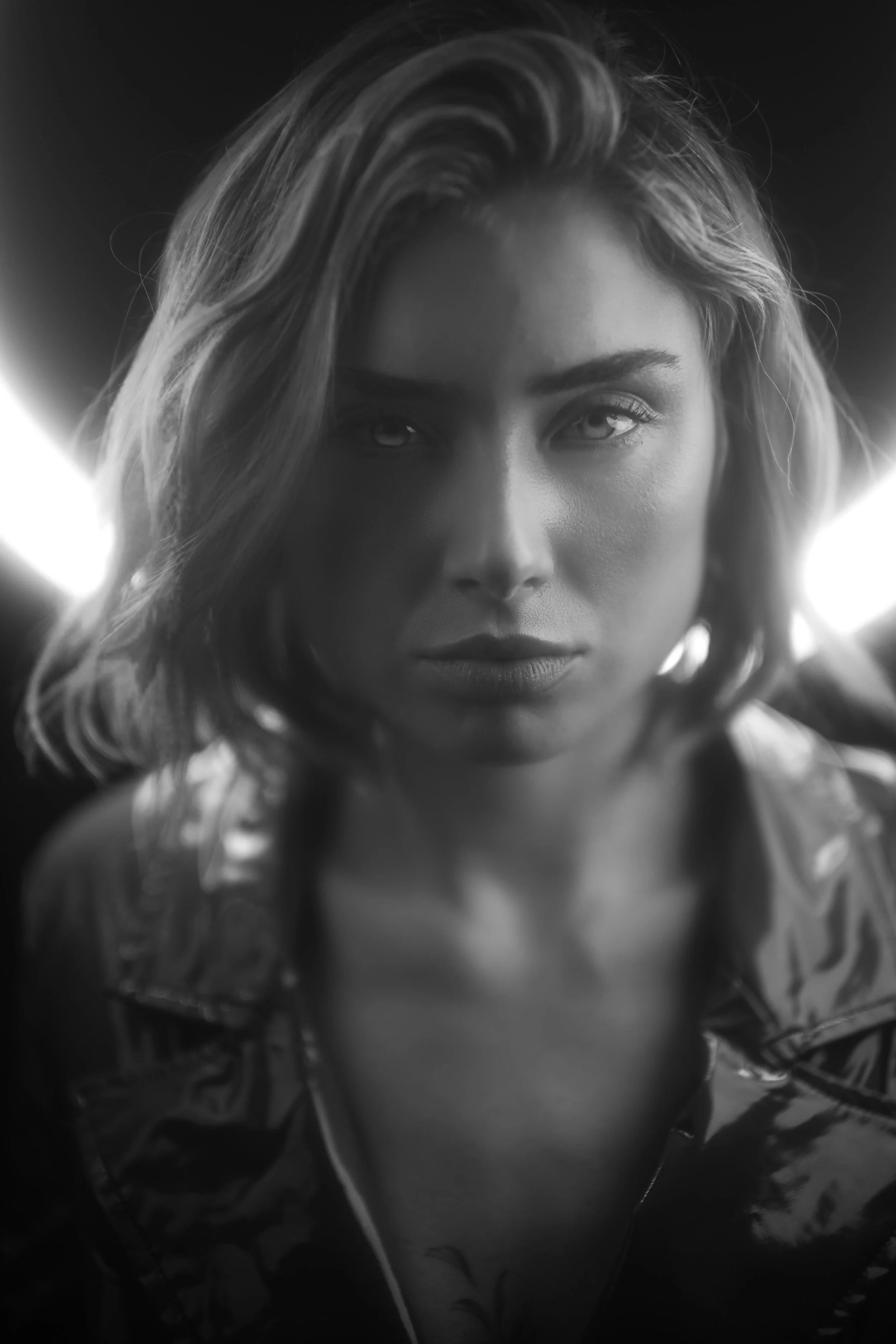 Schwarz-weiß-Fotografie eines jungen Mannes mit kinnlangem Haar, träg Kleidung mit floralem Muster, trägt Ohrhörer, Blick direkt in die Kamera, unscharfer Hintergrund.