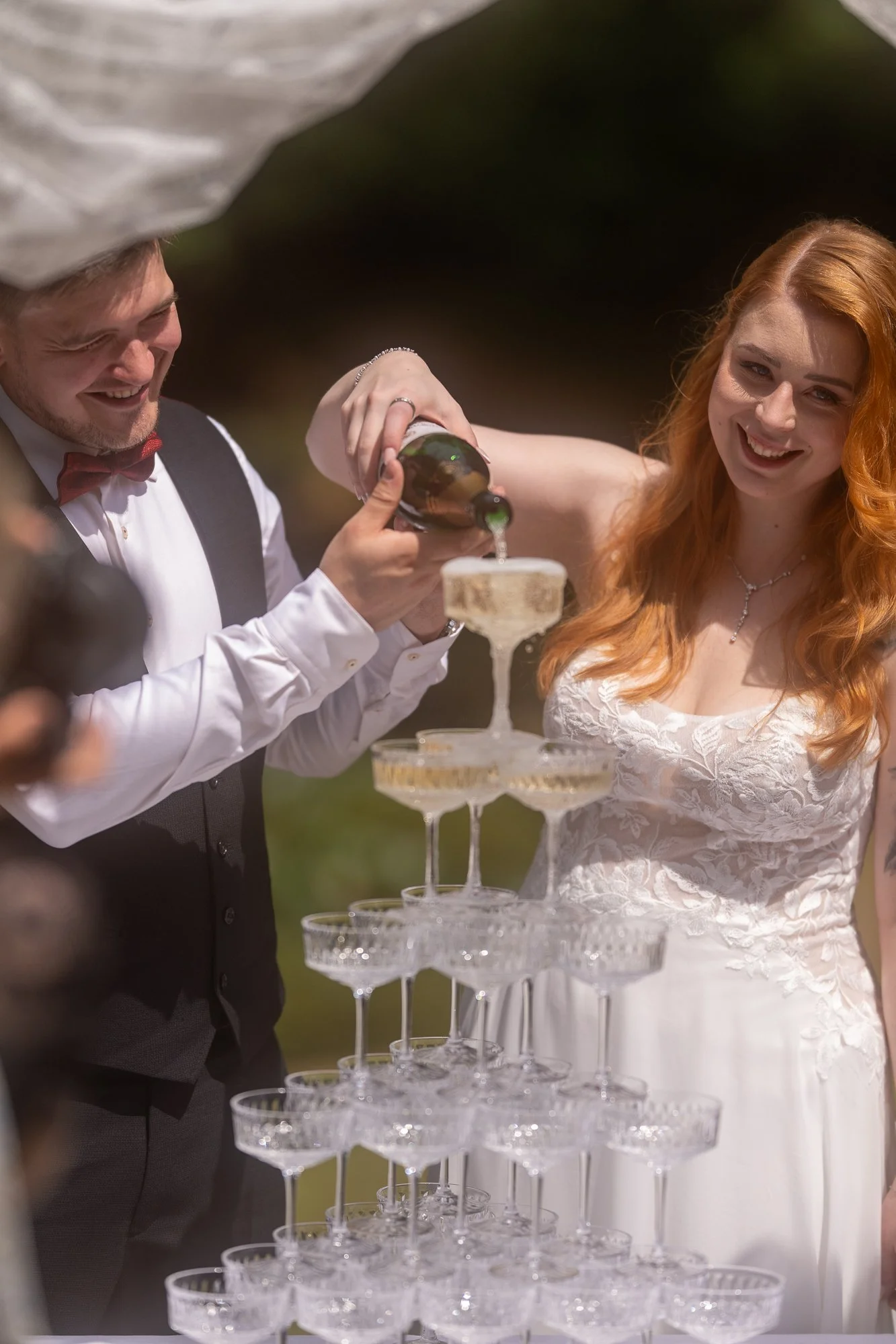 Ein Brautpaar bei einer Hochzeit, das zusammen an einem Champagner-Glückwunsch-Pyramide-Anstoß beteiligt ist, während sie lachend lächeln.