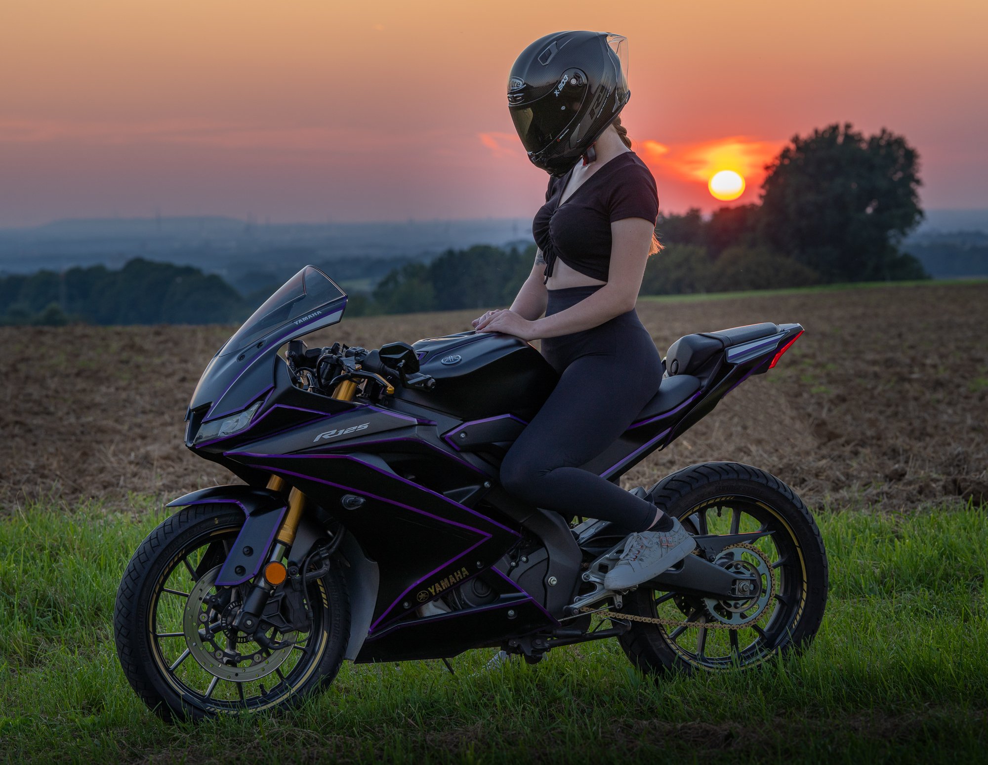 Junge Frau sitzt auf einer schwarzen Yamaha R125 Motorrad, trägt einen schwarzen Helm, eine schwarze Oberbekleidung und graue Turnschuhe, während sie in einem offenen Feld bei Sonnenuntergang steht.