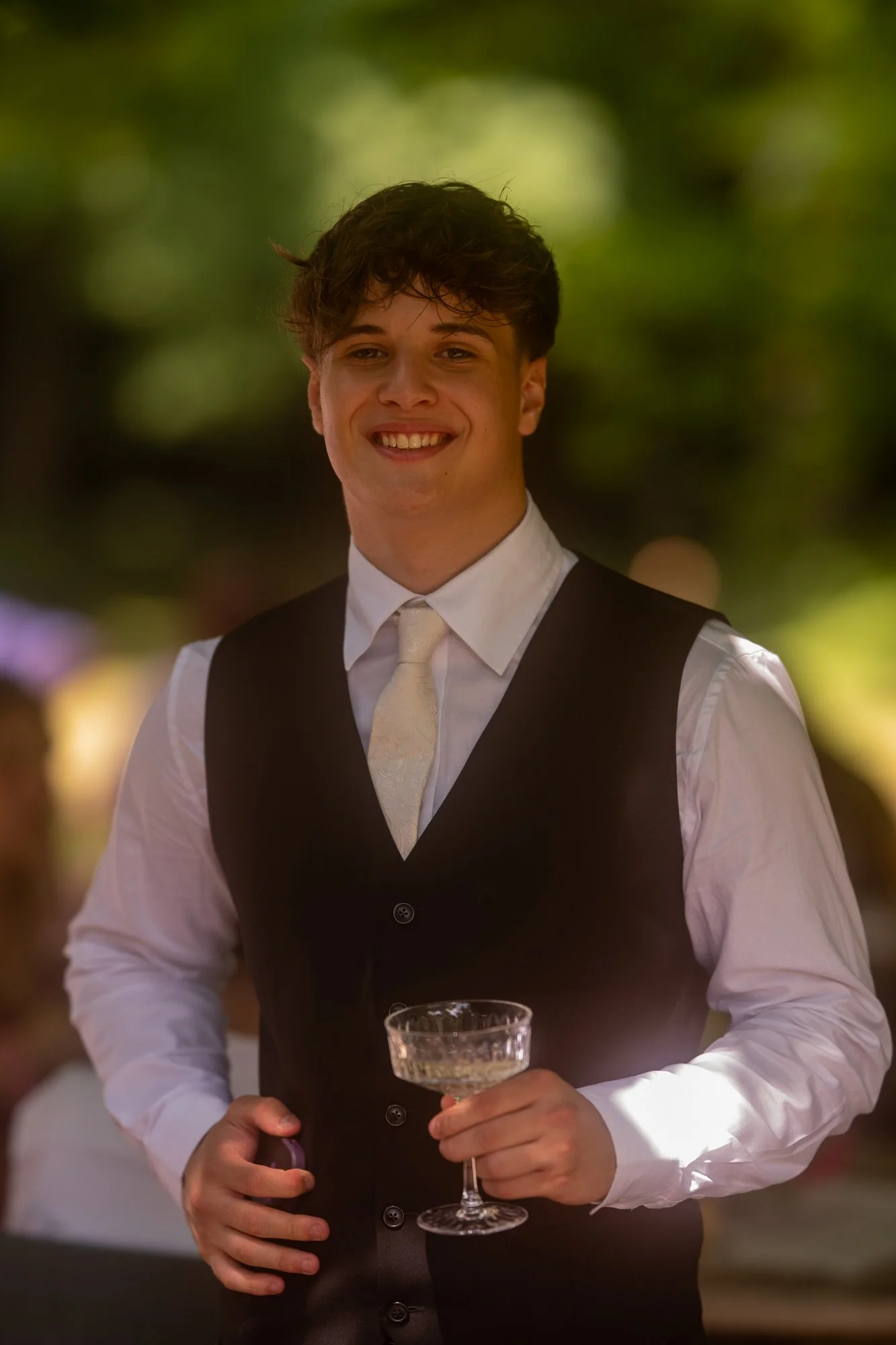 Junger Mann in formeller Kleidung mit Weste und Krawatte, der ein Champagnerglas hält, beim gesellschaftlichen Event.