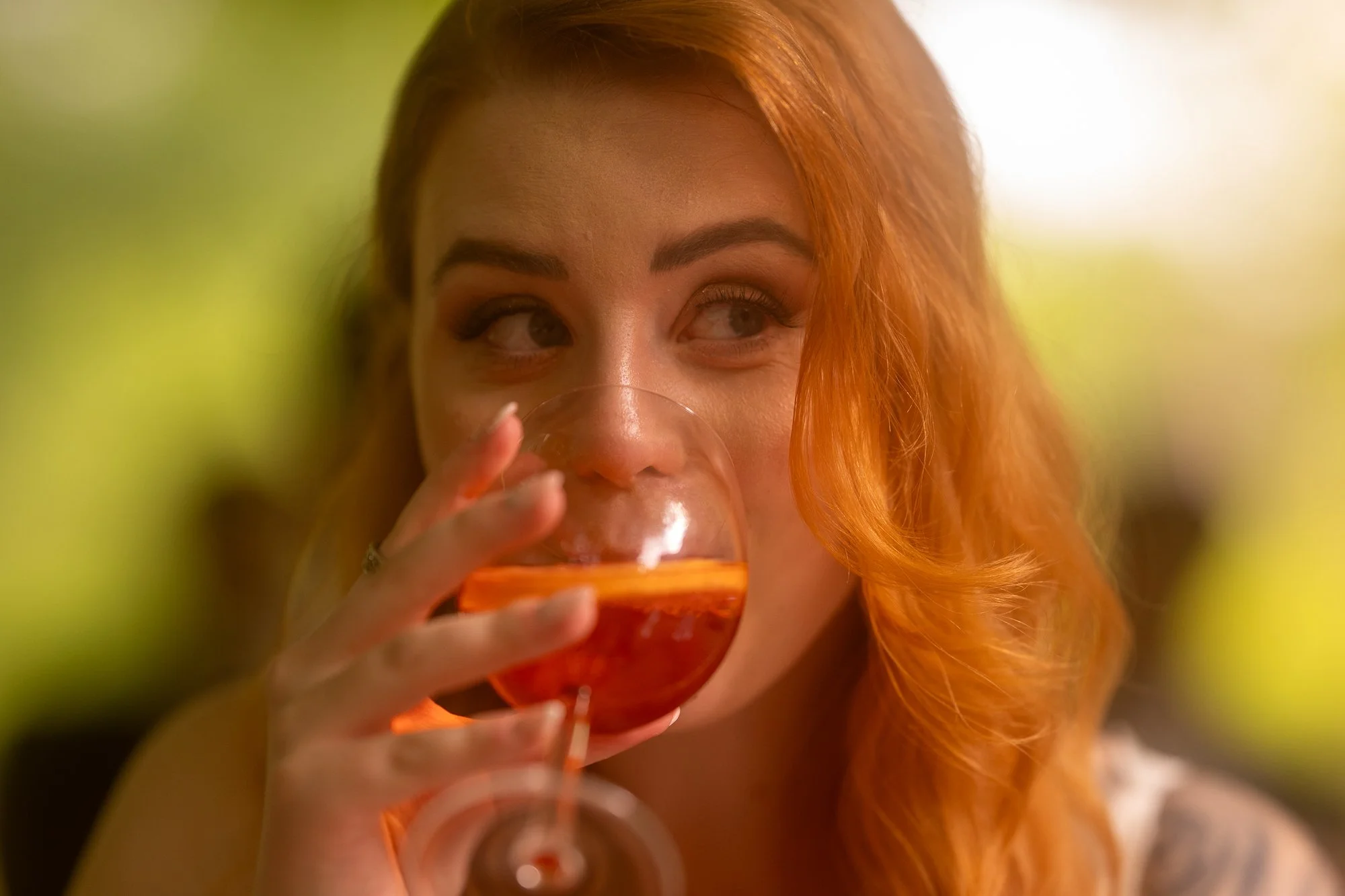 Eine Frau mit roten Haaren trinkt aus einem Glas. Sie schaut seitlich und lächelt leicht.