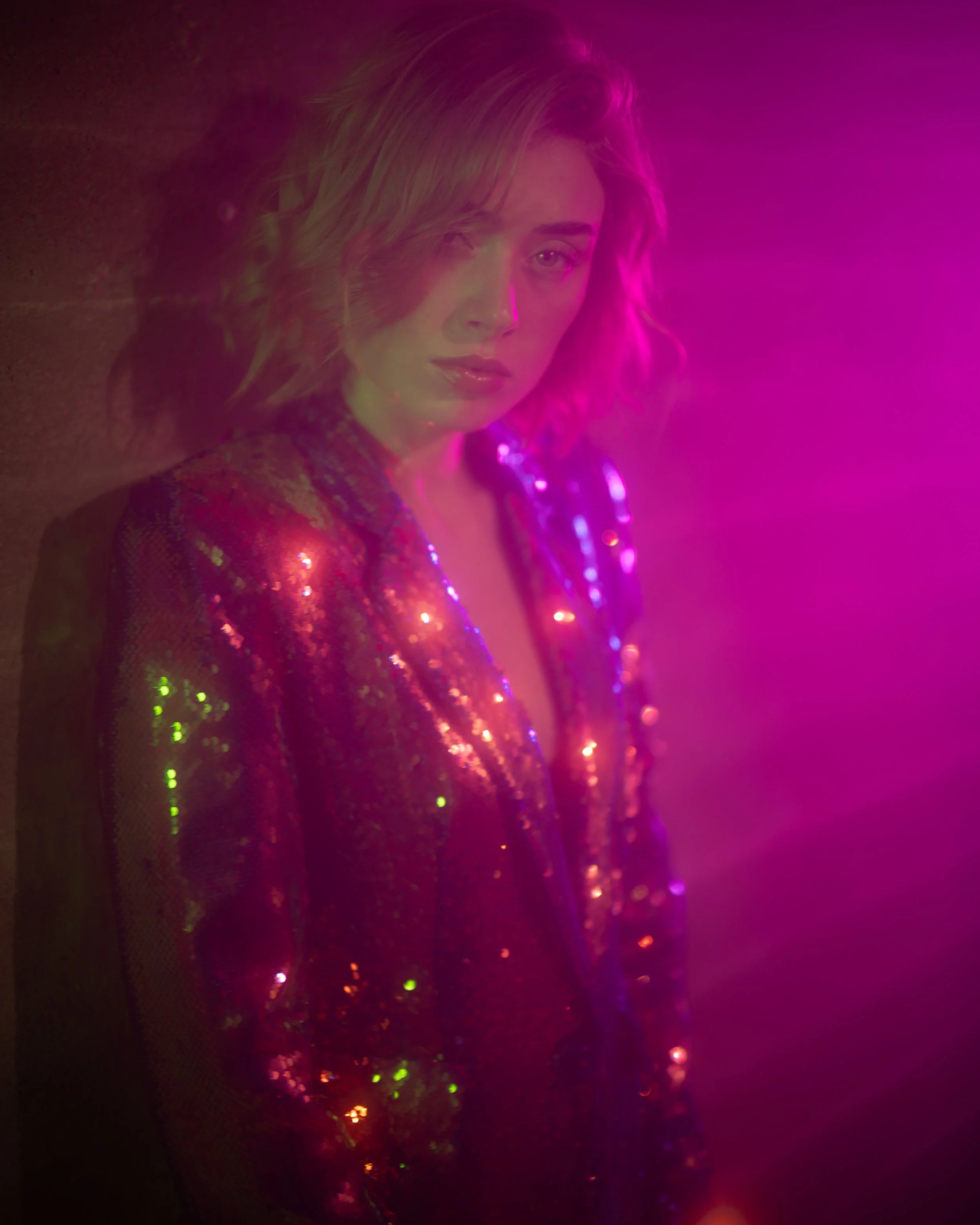 Porträt einer jungen Frau mit lockigem, mittellangem Haar, die auf einem mit bunten Lichtern beleuchteten Hintergrund posiert.