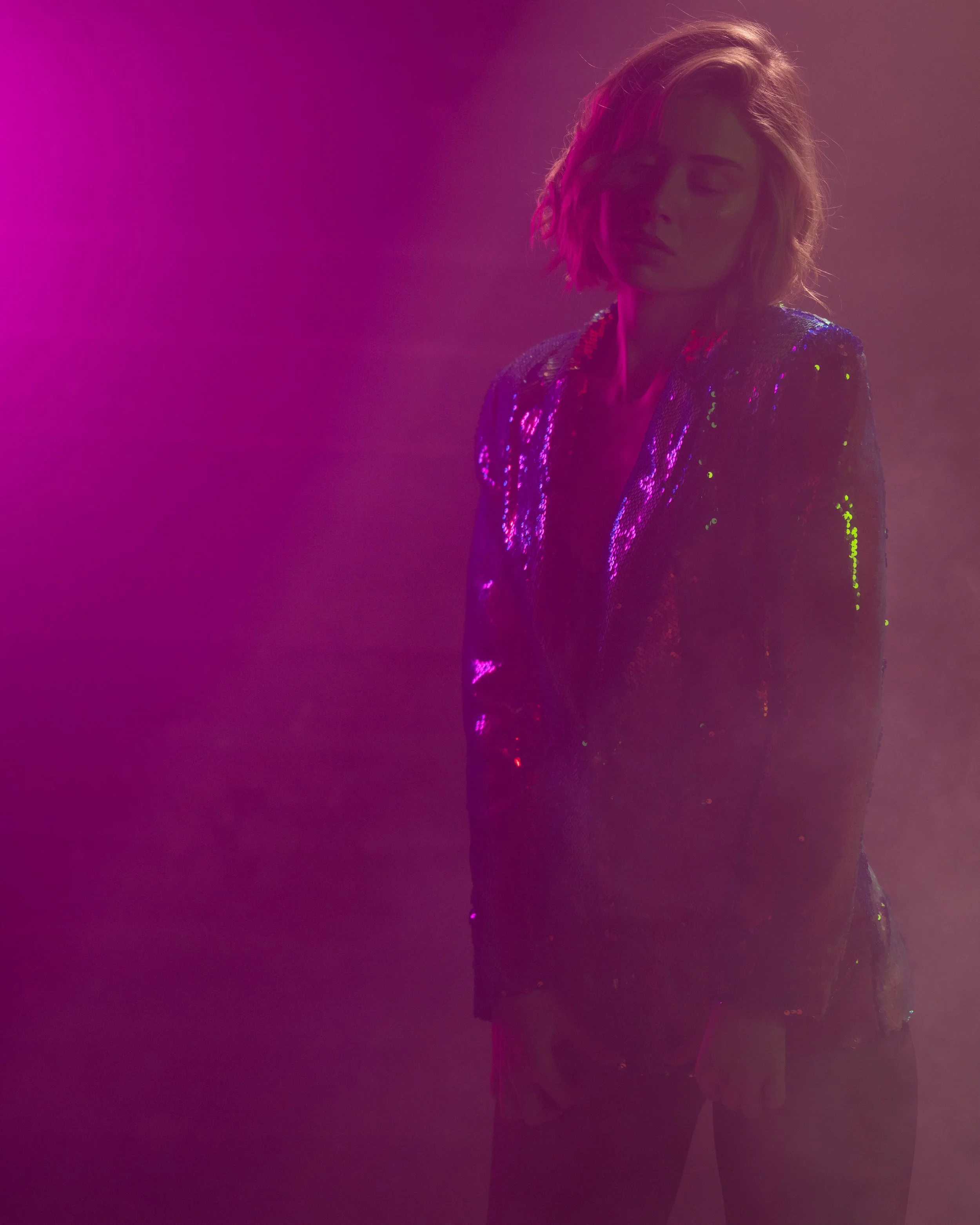 Eine Frau mit welligem Haar trägt eine glänzende, elegante Jacke und steht vor einem bunten, durchscheinenden Licht- und Nebeleffekt.