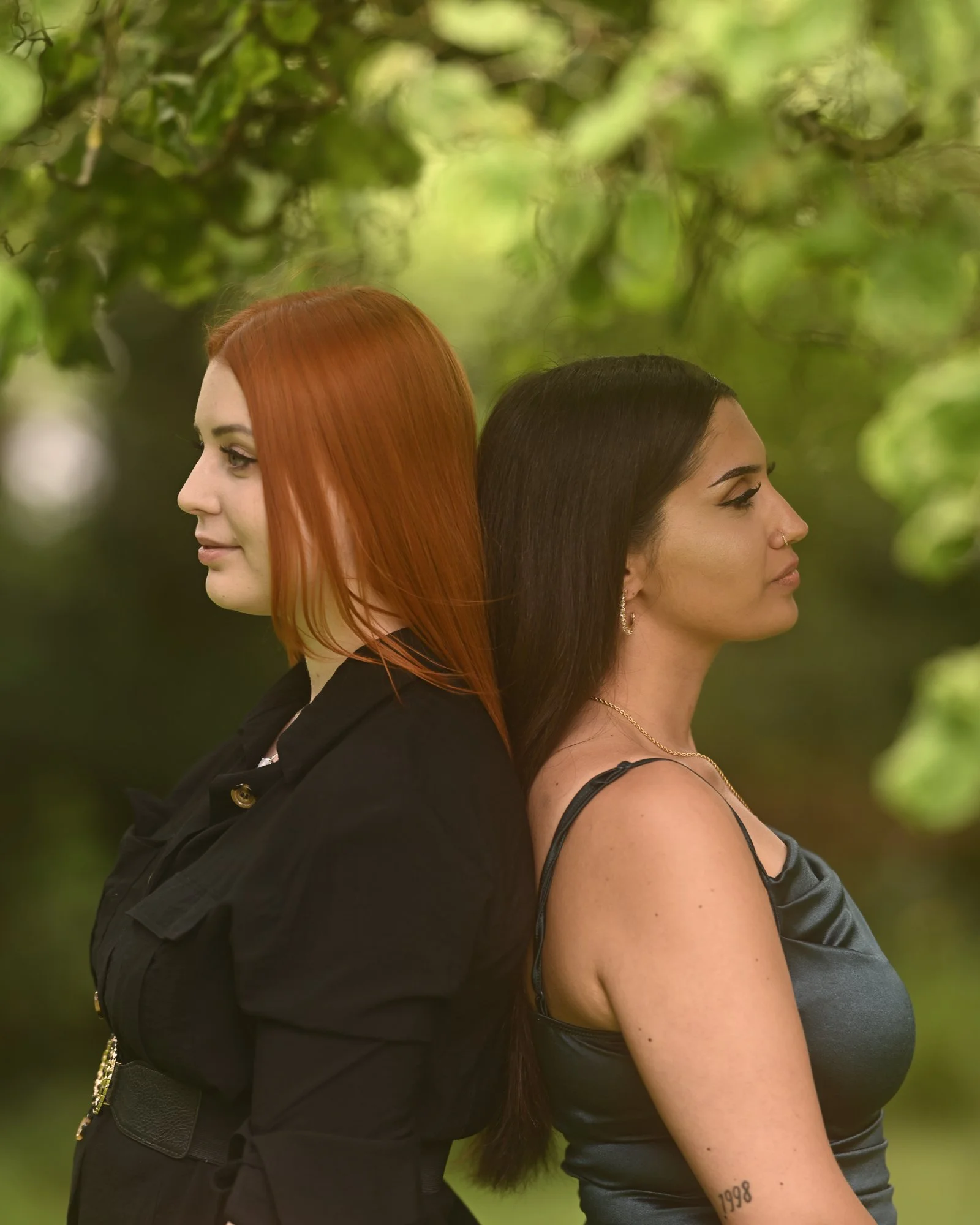 Zwei Frauen stehen Rücken an Rücken in der Natur, eine mit roten, die andere mit schwarzen Haaren, beide sind in Schwarz gekleidet.