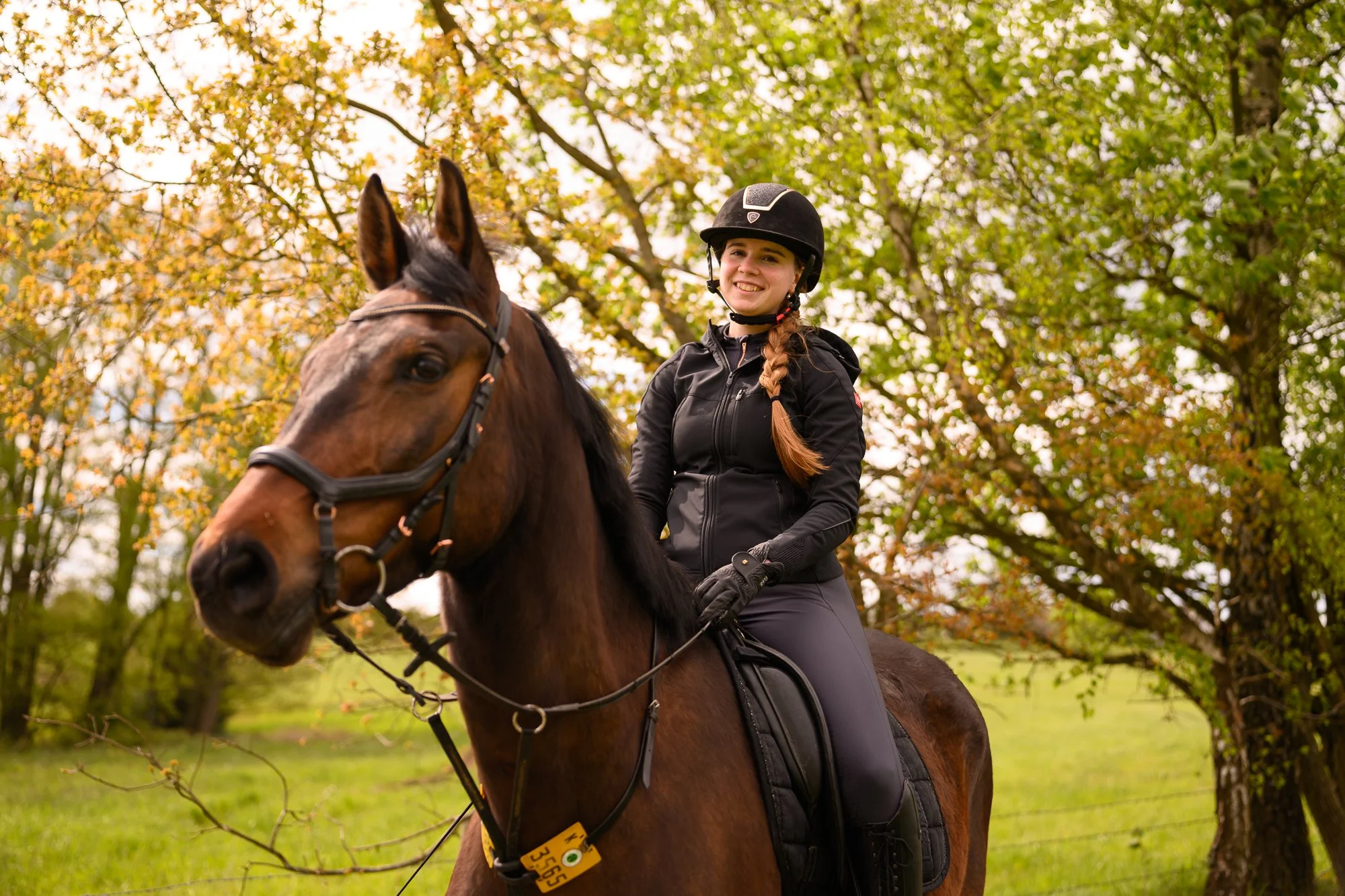 Mädchen in schwarzer Reitjacke trägt Reithelm, reitet auf braunem Pferd durch einen Park mit Herbstlaub, im Hintergrund viele grüne und gelbe Blätter an Bäumen.
