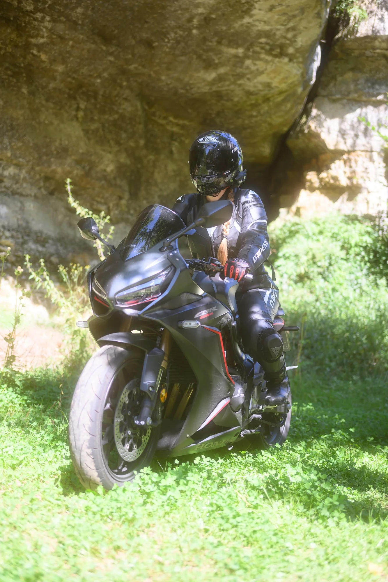 Motorradfahrer auf einer schwarzen Moto-Guzzi V85 TT in der Natur, unter einem Felsvorsprung, umgeben von grünem Gras und Büschen, mit Helm und Schutzkleidung.