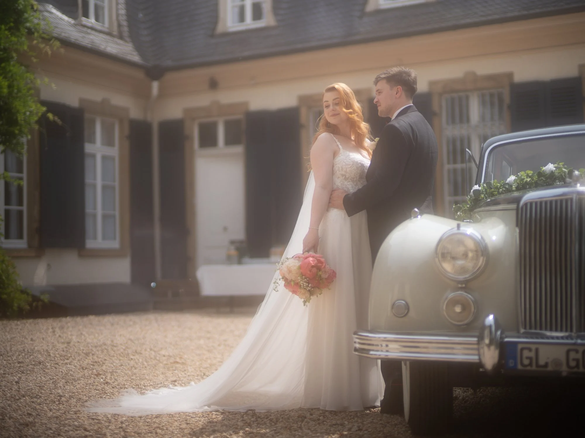 Ein Brautpaar steht neben einem nostalgischen Auto vor einem traditionellen Haus, im Sonnenlicht. Die Braut trägt ein weißes Hochzeitskleid und hält einen Blumenstrauß, während der Bräutigam einen dunklen Anzug trägt.