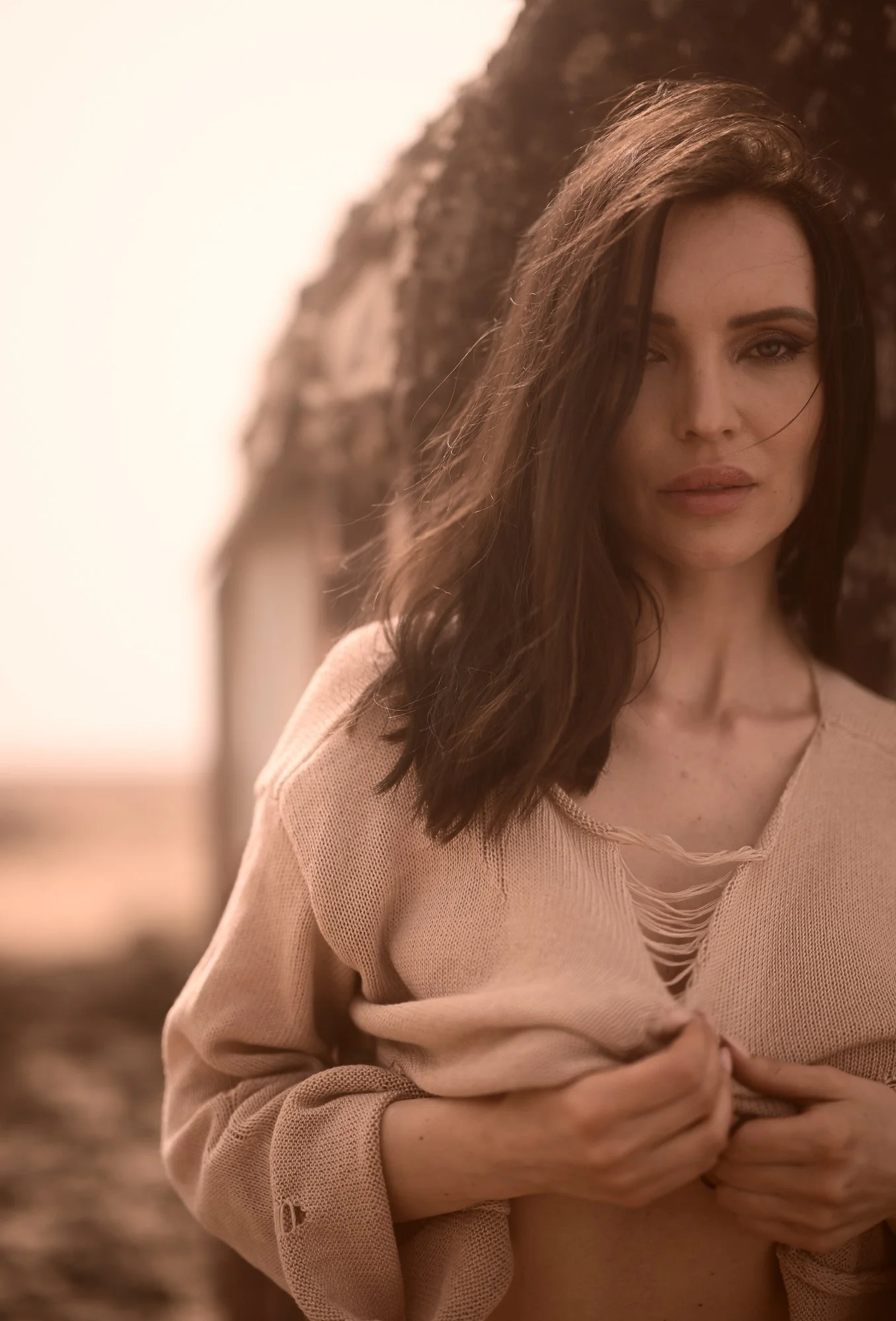 Eine Frau mit braunen, gewellten Haaren und natürlichem Make-up trägt einen beige Pullover mit tiefem Ausschnitt und hält ihre Hände vor ihrem Bauch. Sie steht vor einem Felsen oder einer Wand und ist beim Blick in die Kamera. Das Bild hat eine warme