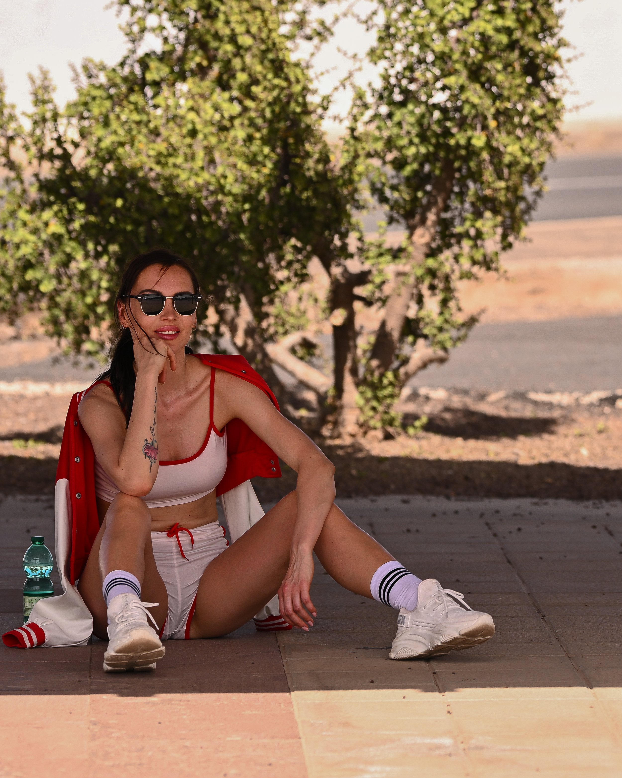 Eine junge Frau in Sportswear mit Sonnenbrille sitzt auf dem Boden, die Beine ausgestreckt, neben einer Wasserflasche, im Hintergrund Bäume und eine Straße.