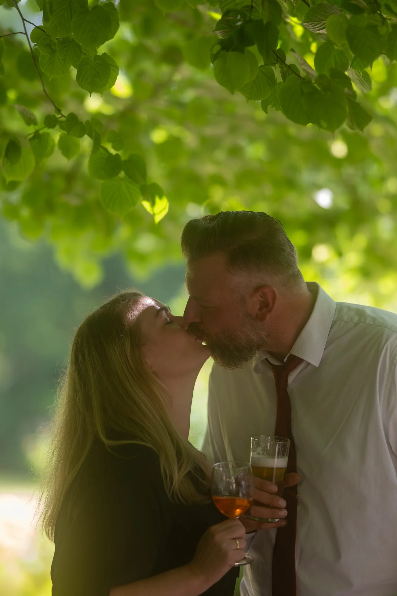 Ein Mann und eine Frau küssen sich unter einem Baum mit grünen Blättern, während sie Wein trinken bei Tageslicht im Freien.