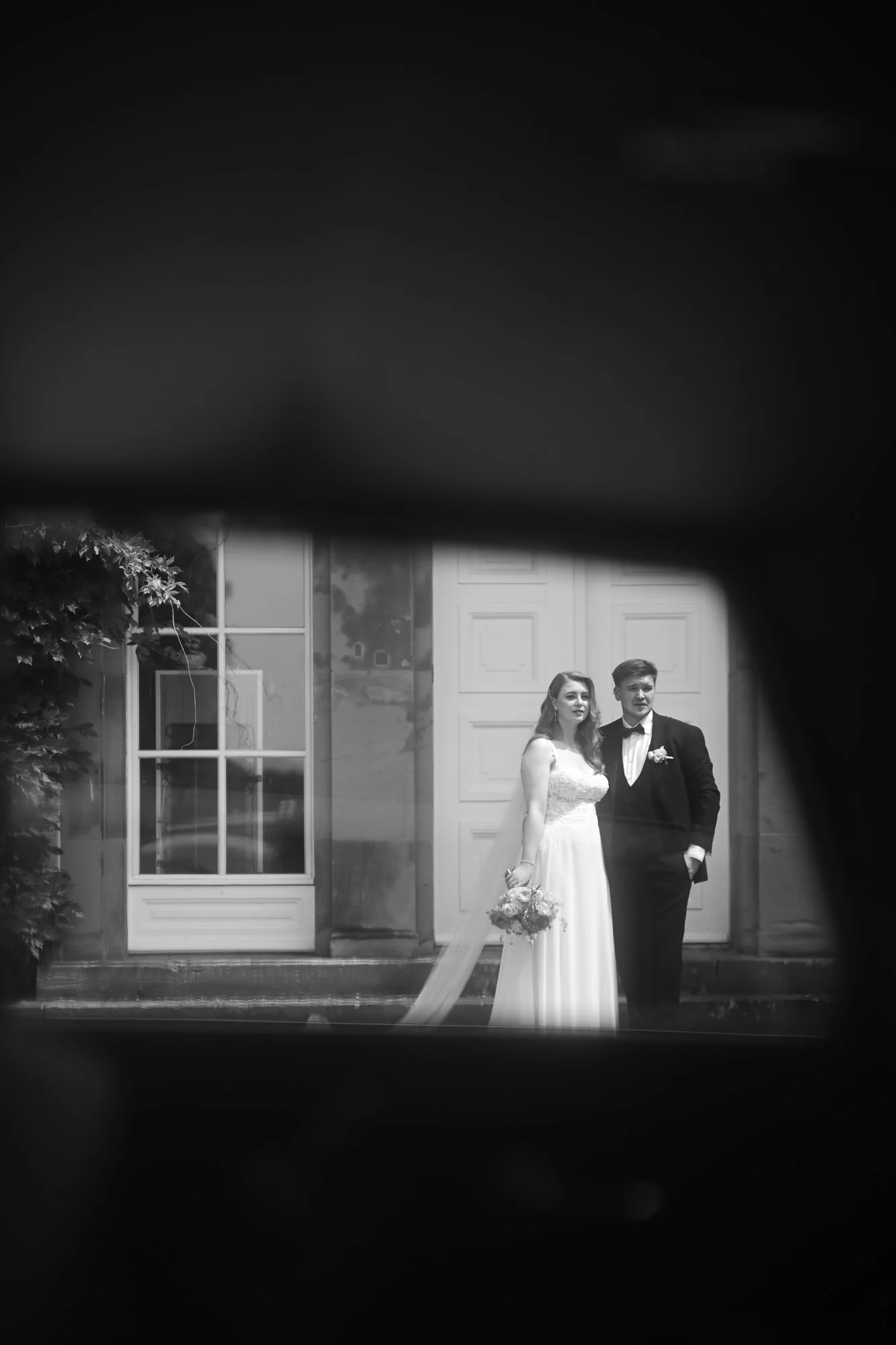 Braut und Bräutigam stehen vor einem Gebäude, vermutlich während einer Hochzeit, im Schwarz-Weiß-Bild.