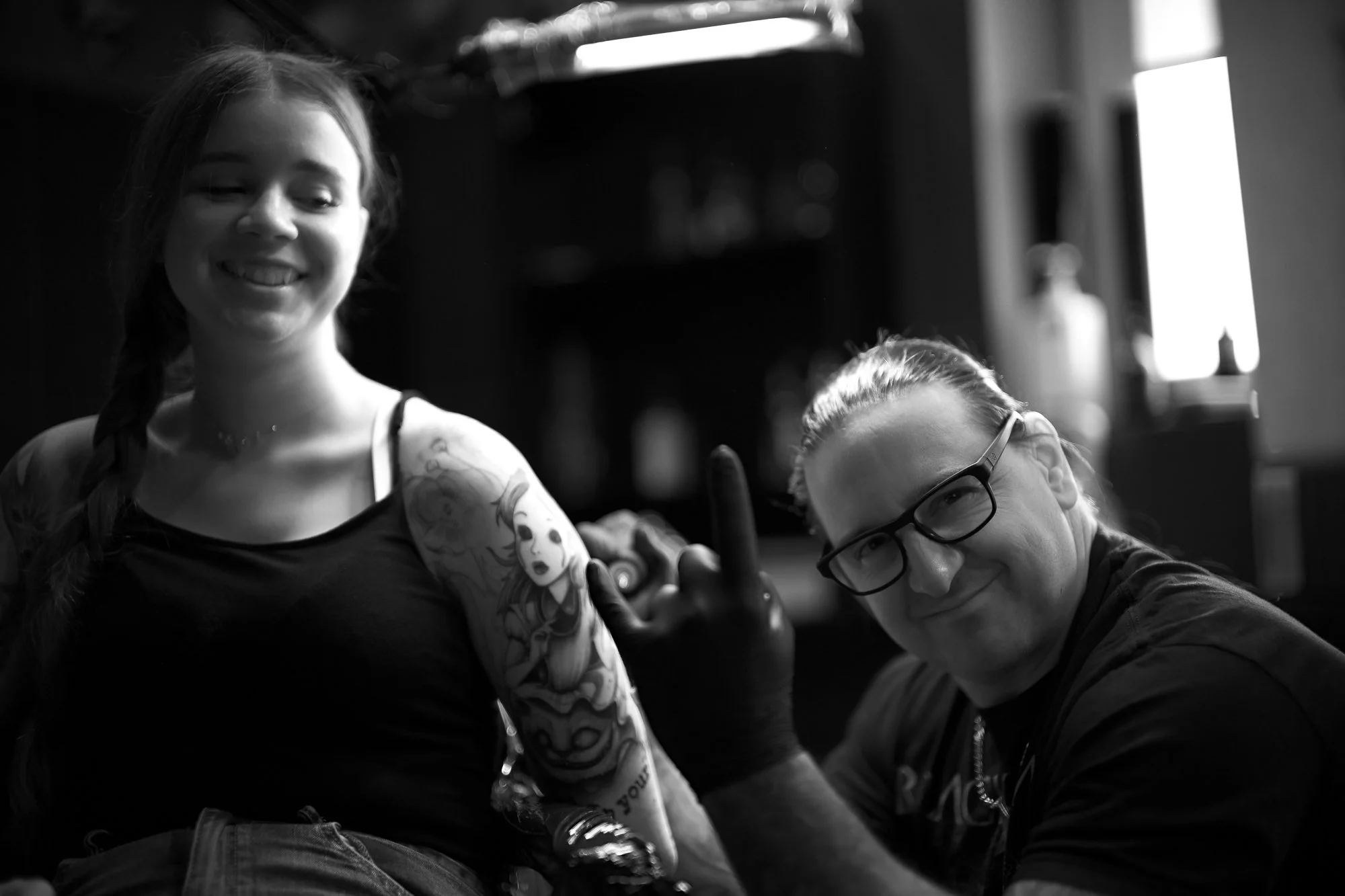 Zwei Menschen posieren in einem Raum, einer mit Tattoos auf dem Arm, der andere mit Brille, beide lächeln und zeigen den Daumen hoch.
