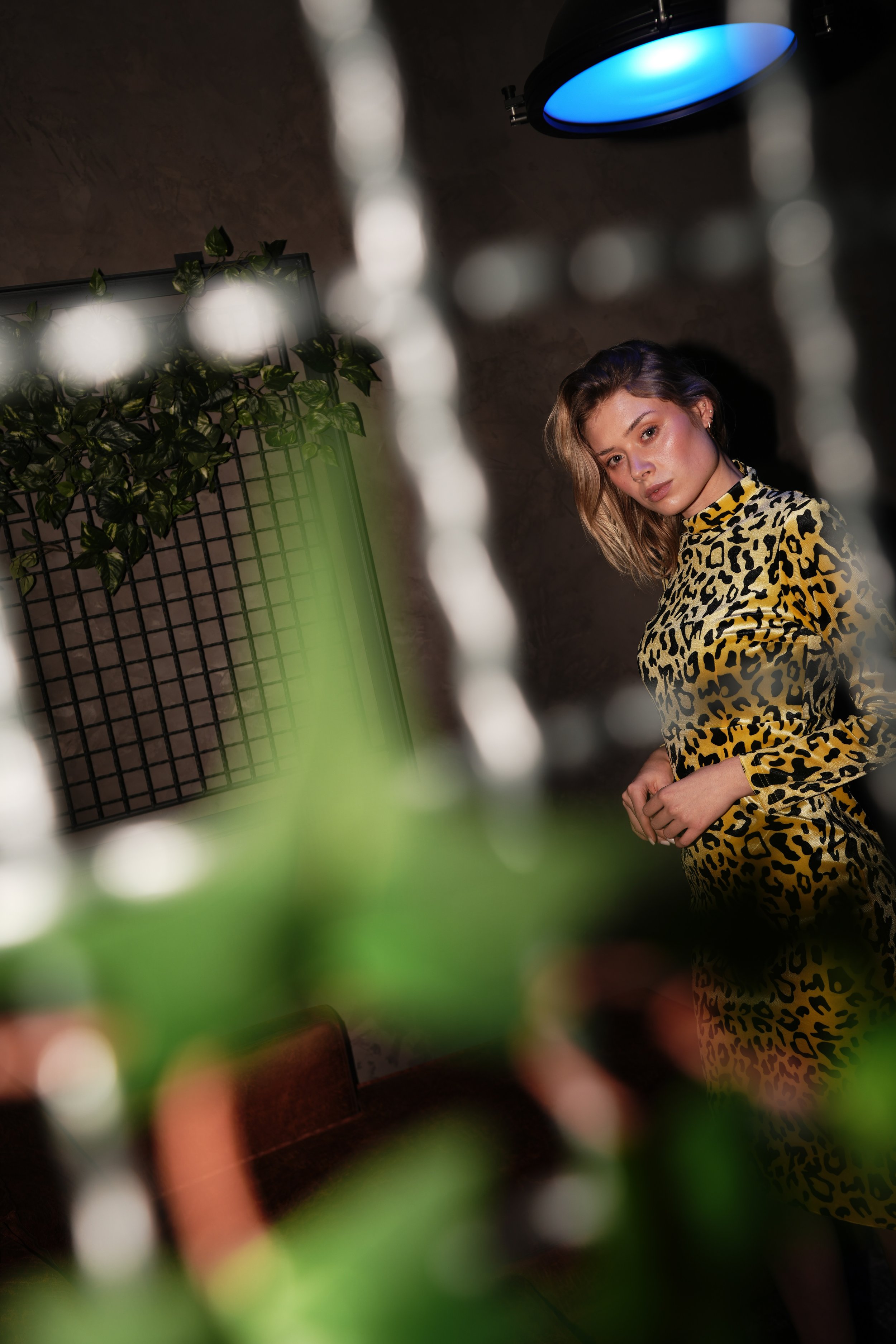 Junge Frau mit herausforderndem Blick trägt eine gelb-schwarz leo-Print Bluse, im Hintergrund eine Pflanzenwand und eine Deckenlampe mit blauer Beleuchtung, unscharf im Vordergrund sind grüne Blätter.