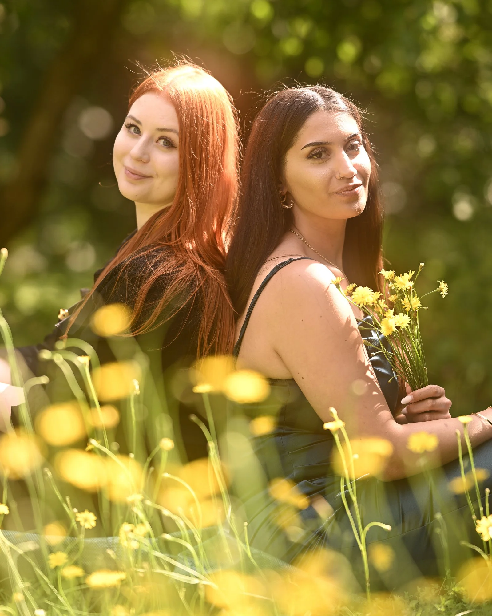 Zwei junge Frauen stehen Rücken an Rücken in einem Blumenfeld mit gelben Blumen, lachend und zufrieden, bei Sonnenlicht, im Freien, mit einem Baum im Hintergrund.
