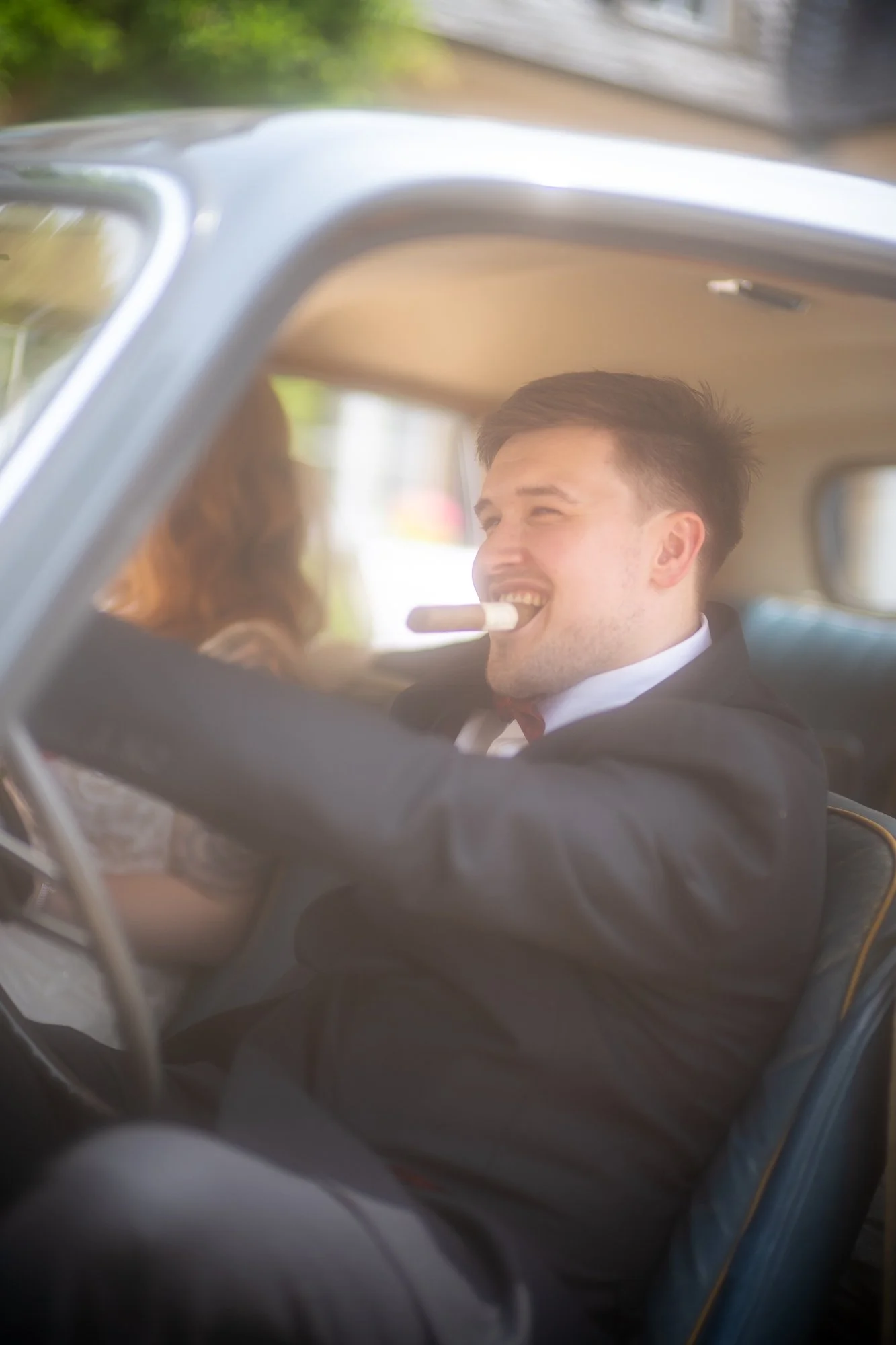 Ein Mann in einem Anzug sitzt in einem Auto und raucht eine Zigarre, während er lächelt. Das Bild ist unscharf und wird durch die Autofenster aufgenommen.