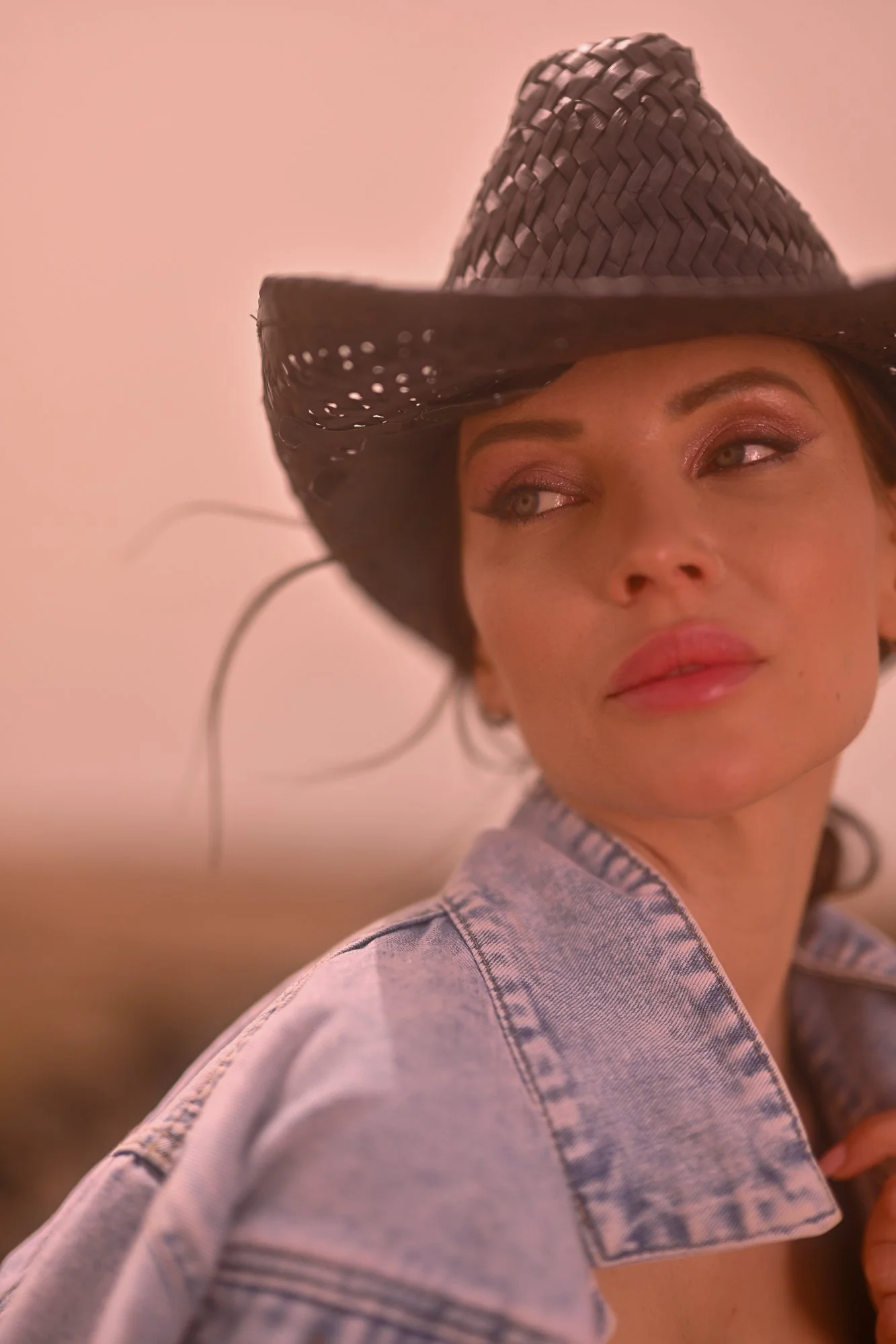 Eine Frau trägt einen schwarzen Strohhut und eine Jeansjacke, blickt seitlich und lächelt leicht.