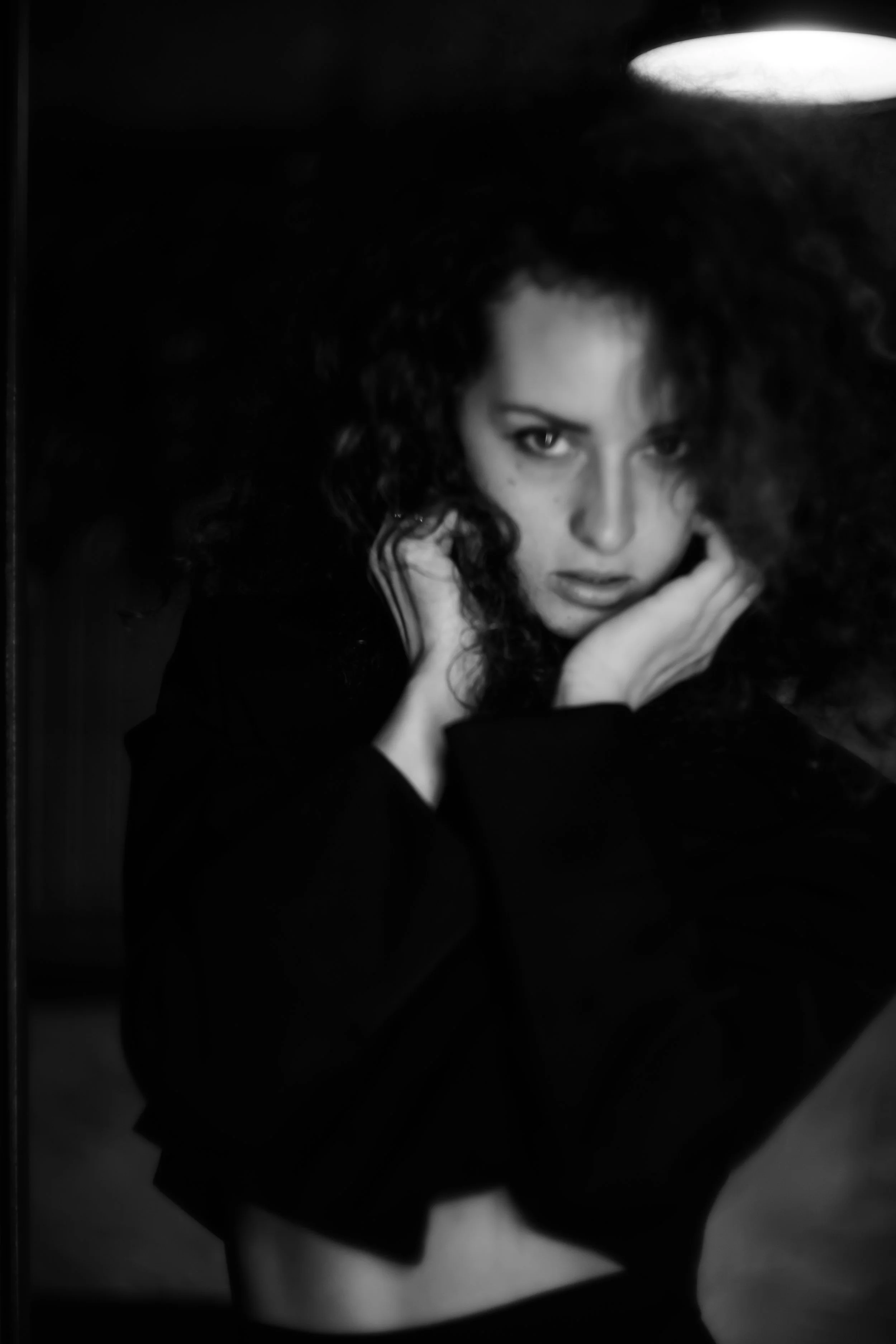 Schwarz-weiß-Fotografie eines jungen Mädchens mit lockigem Haar, das mit den Händen an den Ohren greift und direkt in die Kamera schaut.