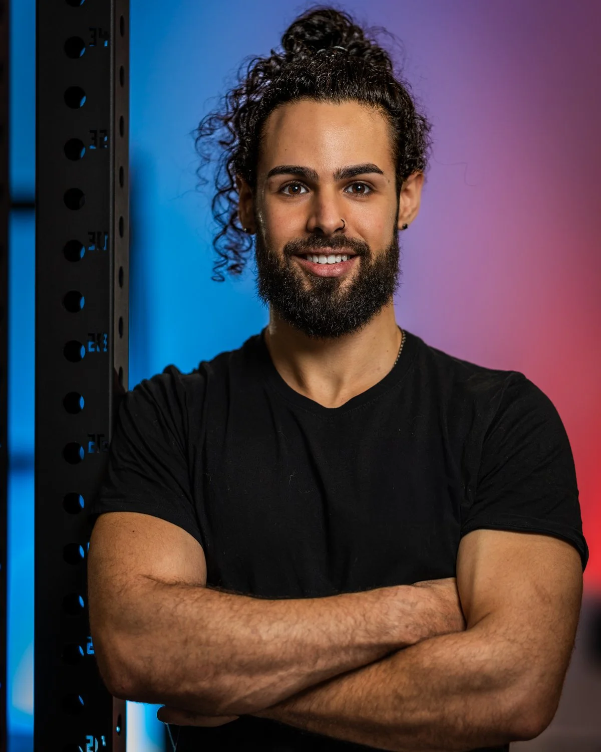 Ein lächelnder Mann mit dunklem T-Shirt, gelocktem Bart und lockigem Haar, der die Arme verschränkt, vor einem grauen und blauen Hintergrund in einem Fitnessstudio oder ähnlichem Raum.