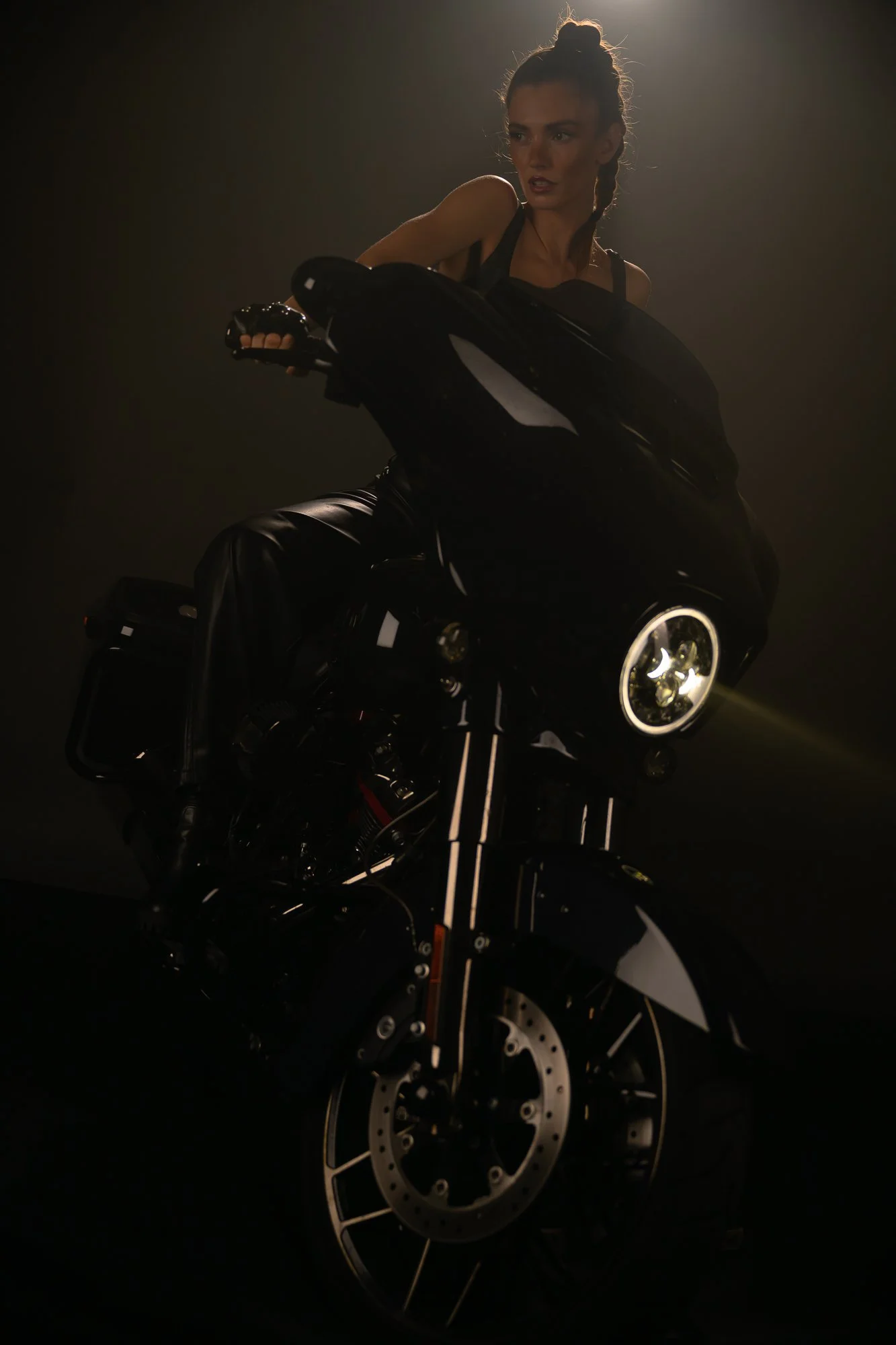 Eine Frau sitzt auf einem schwarzen Motorrad, das in einem dunklen Raum steht, mit Seitenlichtrichtung, der Kopf nach rechts, oben, die Mittelung leuchtet. Die Frau trägt ein schwarzes Top und Jeans, hat ihr Haar in einem Dutt.