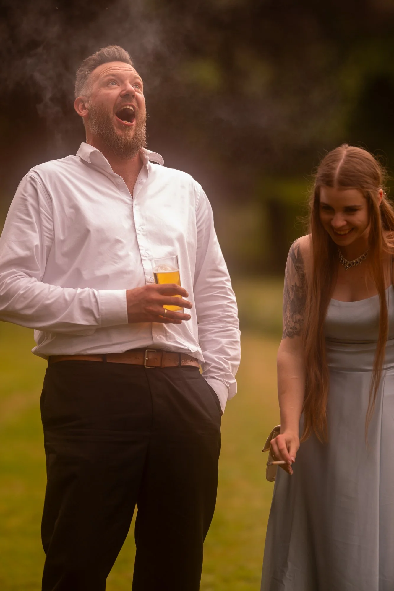 Zwei Personen, ein Mann mit einem weißen Hemd, der einen drink in der Hand hält, und eine Frau mit langen roten Haaren und einem leichten Kleid, lachen und lachen im Freien bei Sonnenuntergang.