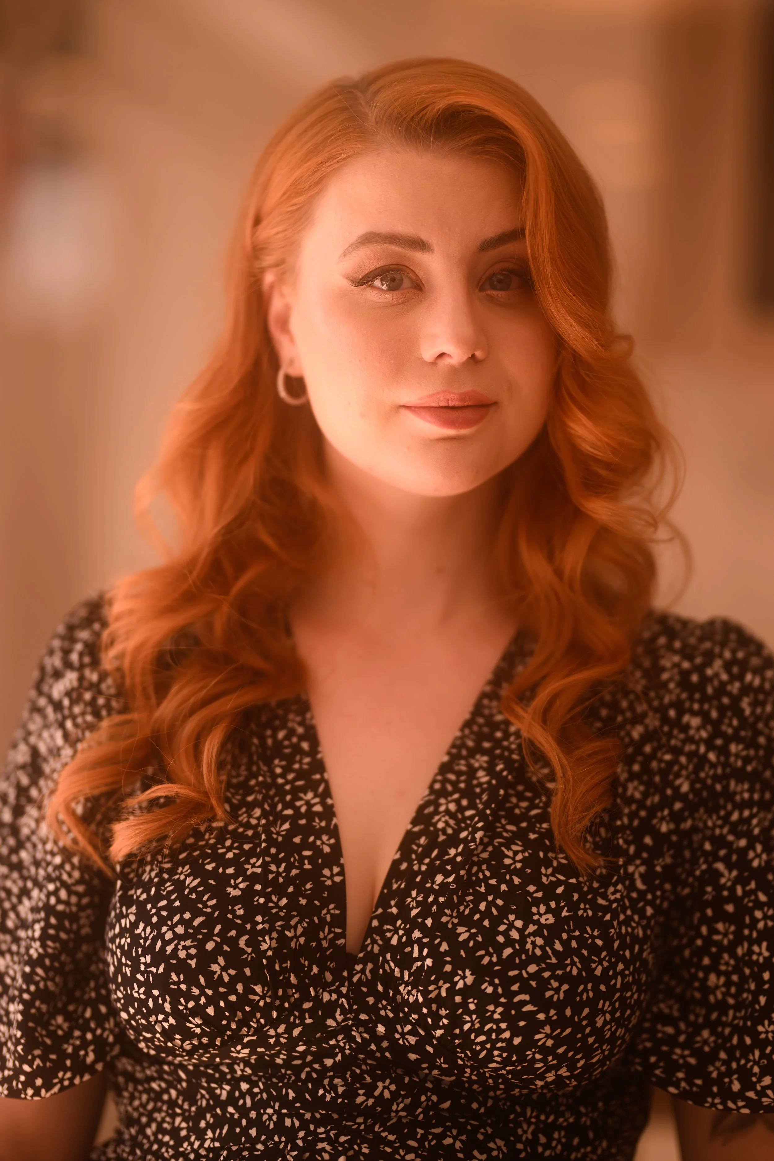 Ein Porträt einer Frau mit langen, lockigen roten Haaren, die auf einem dunkel gemusterten Kleid mit V-Ausschnitt steht, vor einem warmen, unscharfen Hintergrund.