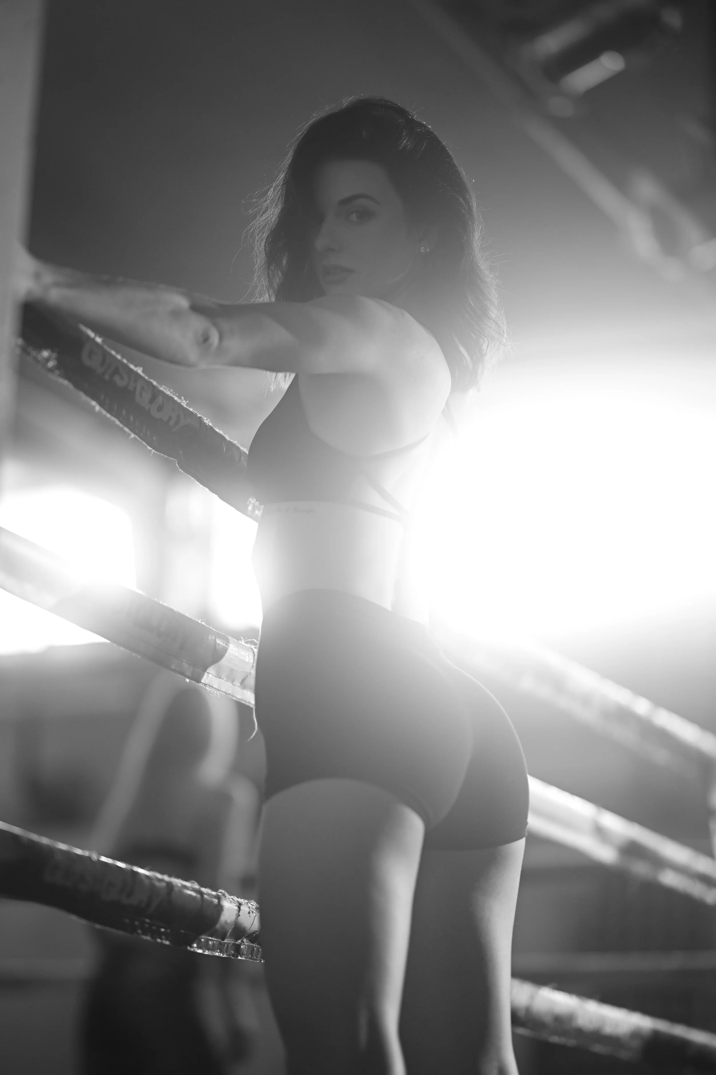 Schwarz-Weiß-Foto einer Boxerin, die im Ring trainiert, mit Fokus auf ihren Oberkörper und Blick, helles Licht im Hintergrund.