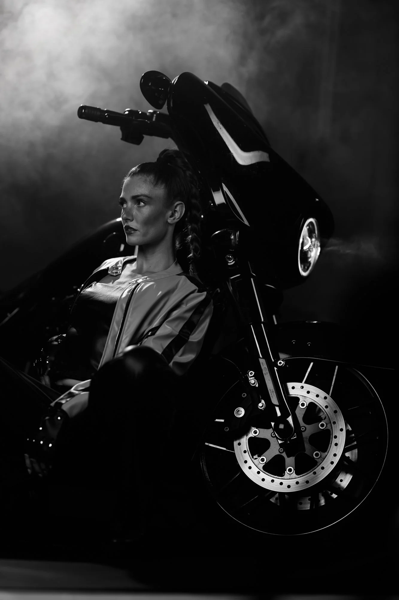 Schwarz-weiß-Fotografie eines jungen Mannes mit kurzen Haaren im Motorradhelm, sitzend auf einem schwarzen Motorrad, mit Glanzlichtern auf der Haut, vor dunklem Hintergrund.