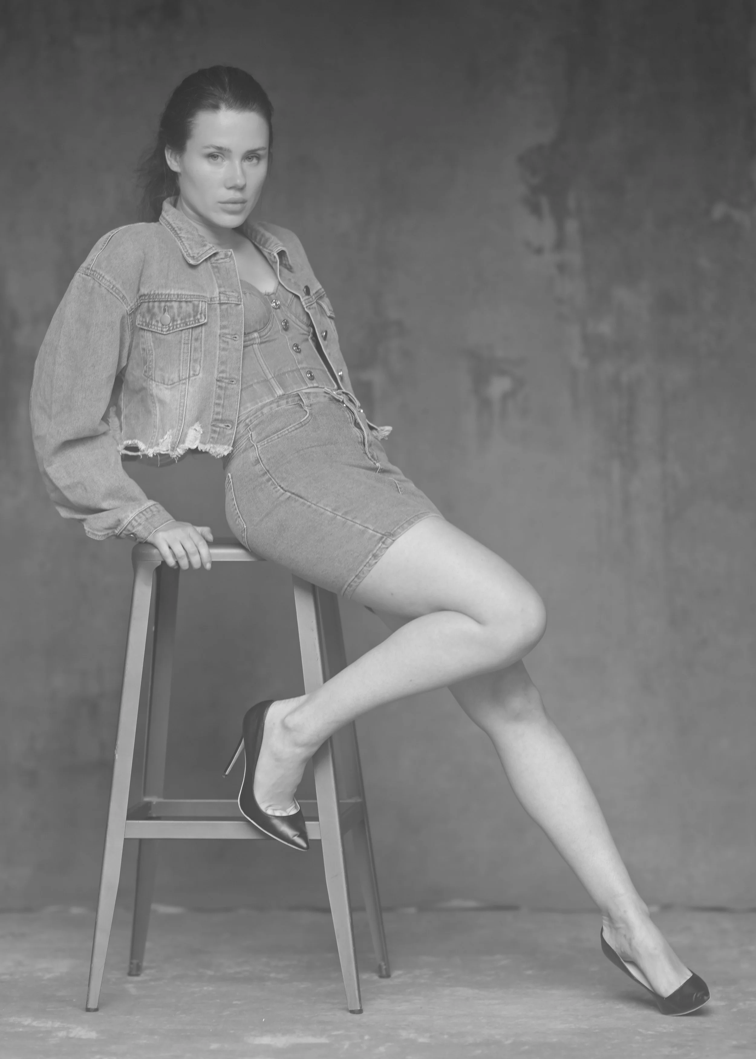 Schwarz-weiß-Foto einer jungen Frau, die auf einem Holzstuhl sitzt. Sie trägt eine Jeansjacke, ein Jeansrock und Schuhe mit hohen Absätzen. Ihr Blick ist nach vorne gerichtet, und die Kulisse ist unscharf.