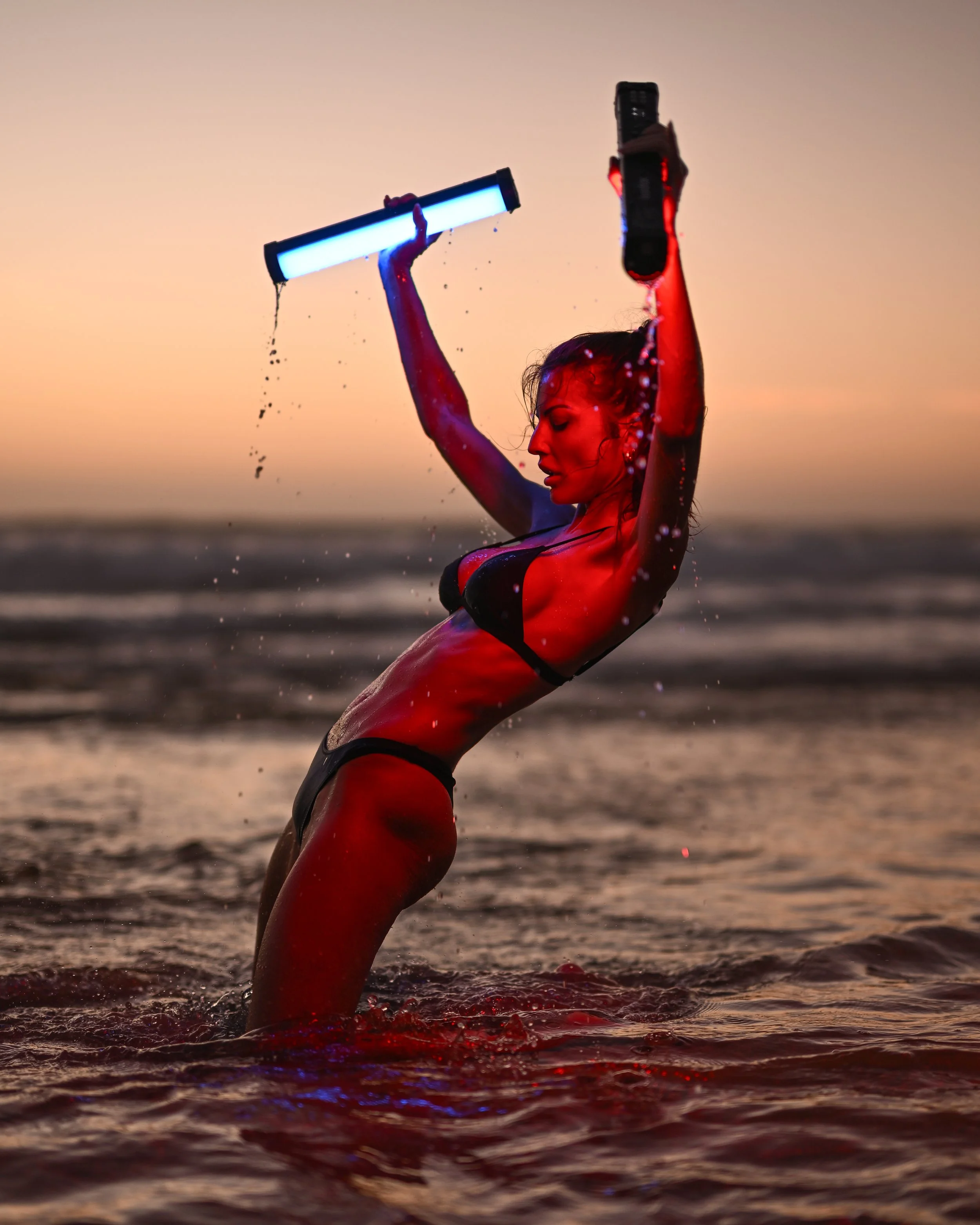 Eine Frau im Bikini beim Wassersport bei Sonnenuntergang am Strand. Sie hält ein beleuchtetes Schwimmgerät in den Händen.