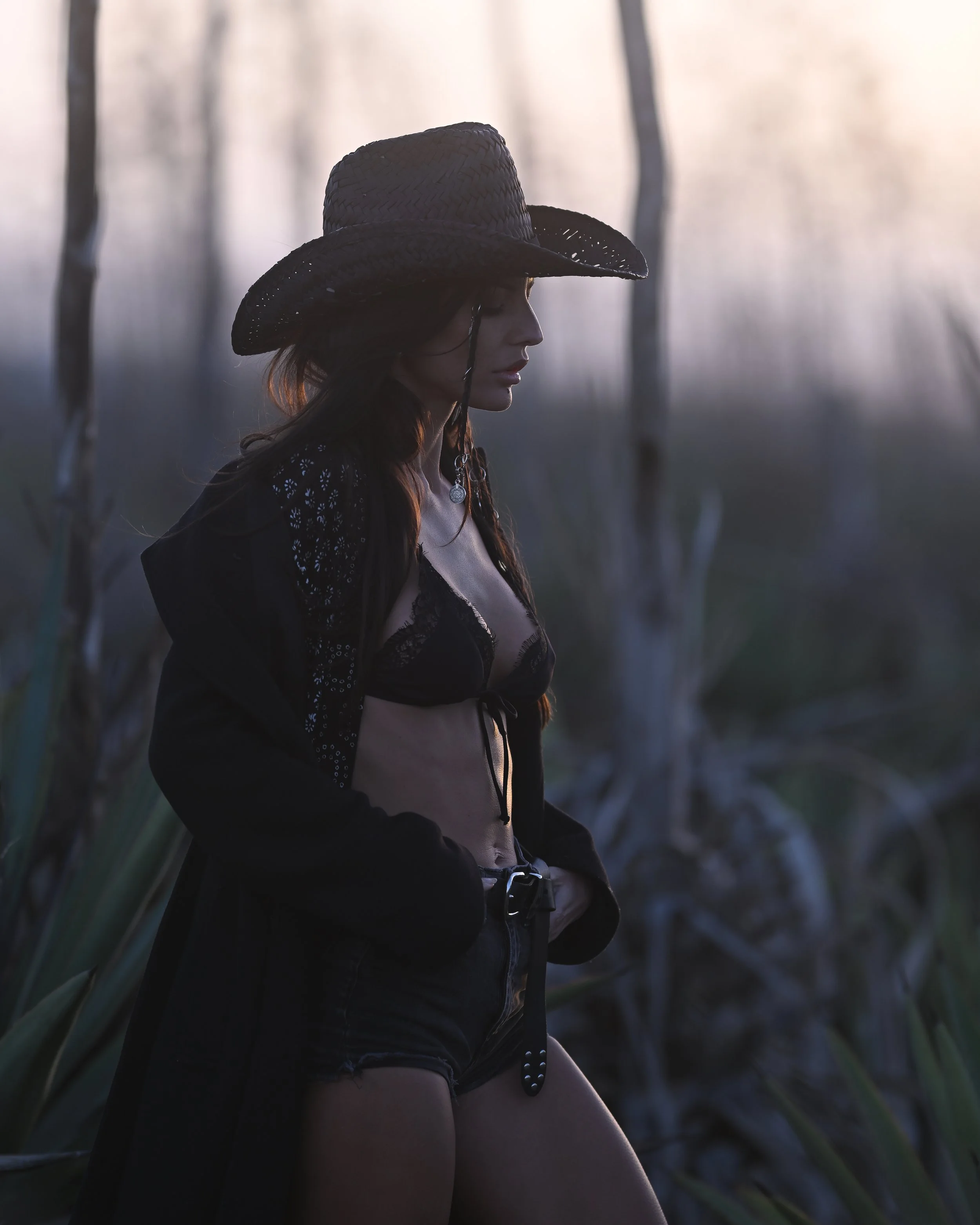 Eine junge Frau trägt einen schwarzen Strohhut und schwarze Kleidung, sitzt neben Pflanzen, bei Sonnenuntergang oder Dämmerung.