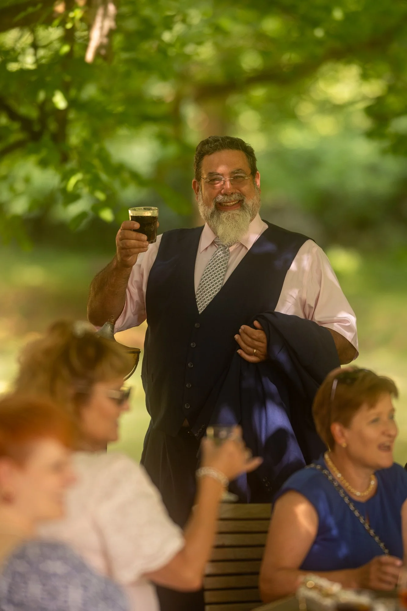 Ein Mann mit Brille und grauem Bart hält ein Glas Bier und lächelt in einem grünen, bewaldeten Hintergrund, während um ihn herum Menschen an einem Tisch sitzen und sprechen.