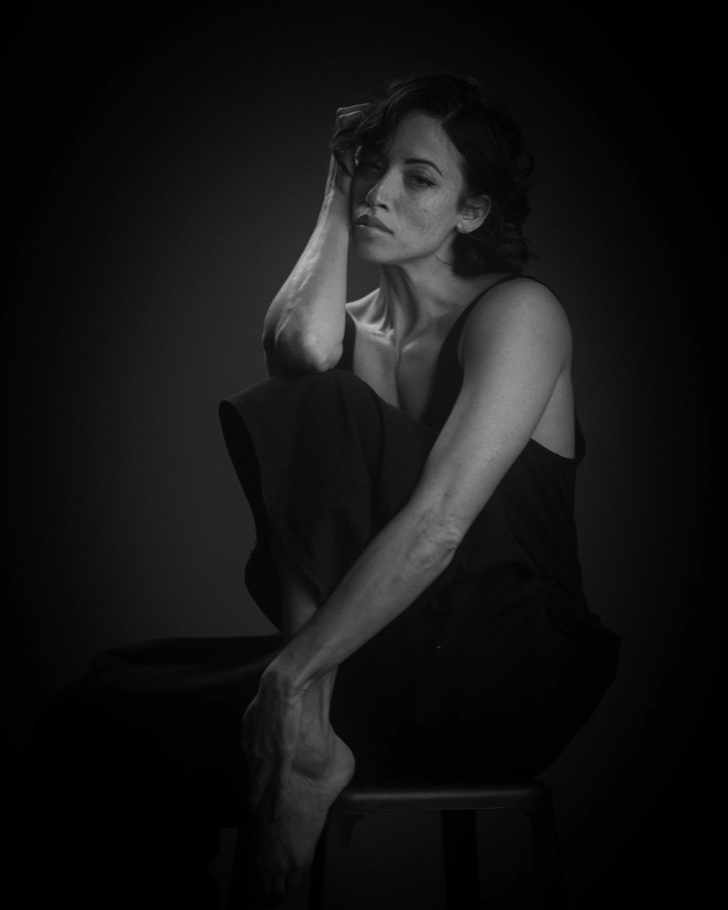 Schwarz-weiß-Foto einer Frau mit dunklen, lockigen Haaren, die ihre Hand auf den Kopf legt. Sie trägt ein dunkles Top und sitzt auf einem Stuhl, vor dunklem Hintergrund.