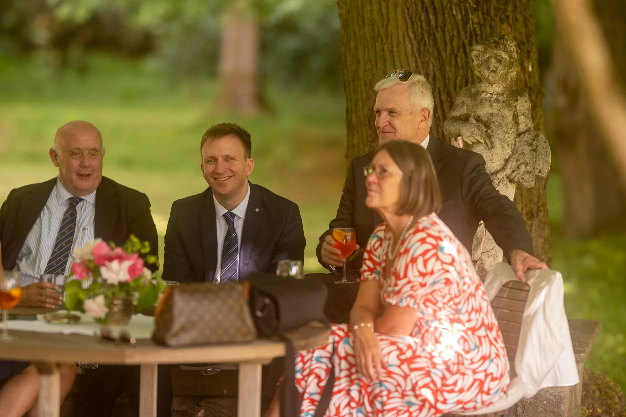 Gruppen von Menschen sitzen bei einem Essen im Freien unter Bäumen, einige lächeln, eine Frau trägt ein rotes und weißes Kleid und hält ein Glas Rotwein.