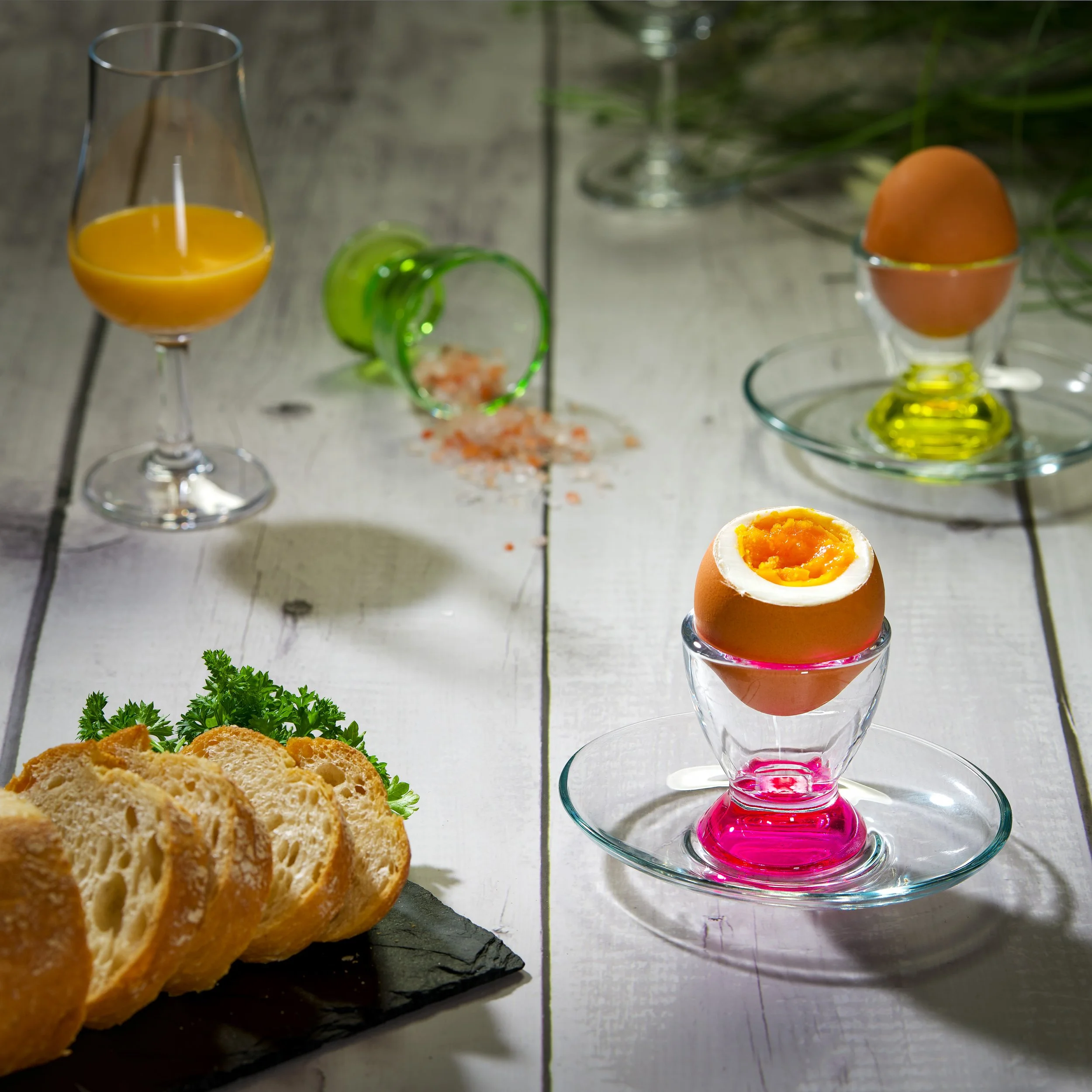 Ein Glas Orangensaft, Eier in einem Eierbecher, Salz- und Pfeffermühle, frische Petersilie, und Knabbereien auf einem Holzbrett auf einem weißen Holztisch. Im Hintergrund eine elegante Vase mit grünen Zweigen, auf einem hellen Tisch. Die Szene ist he